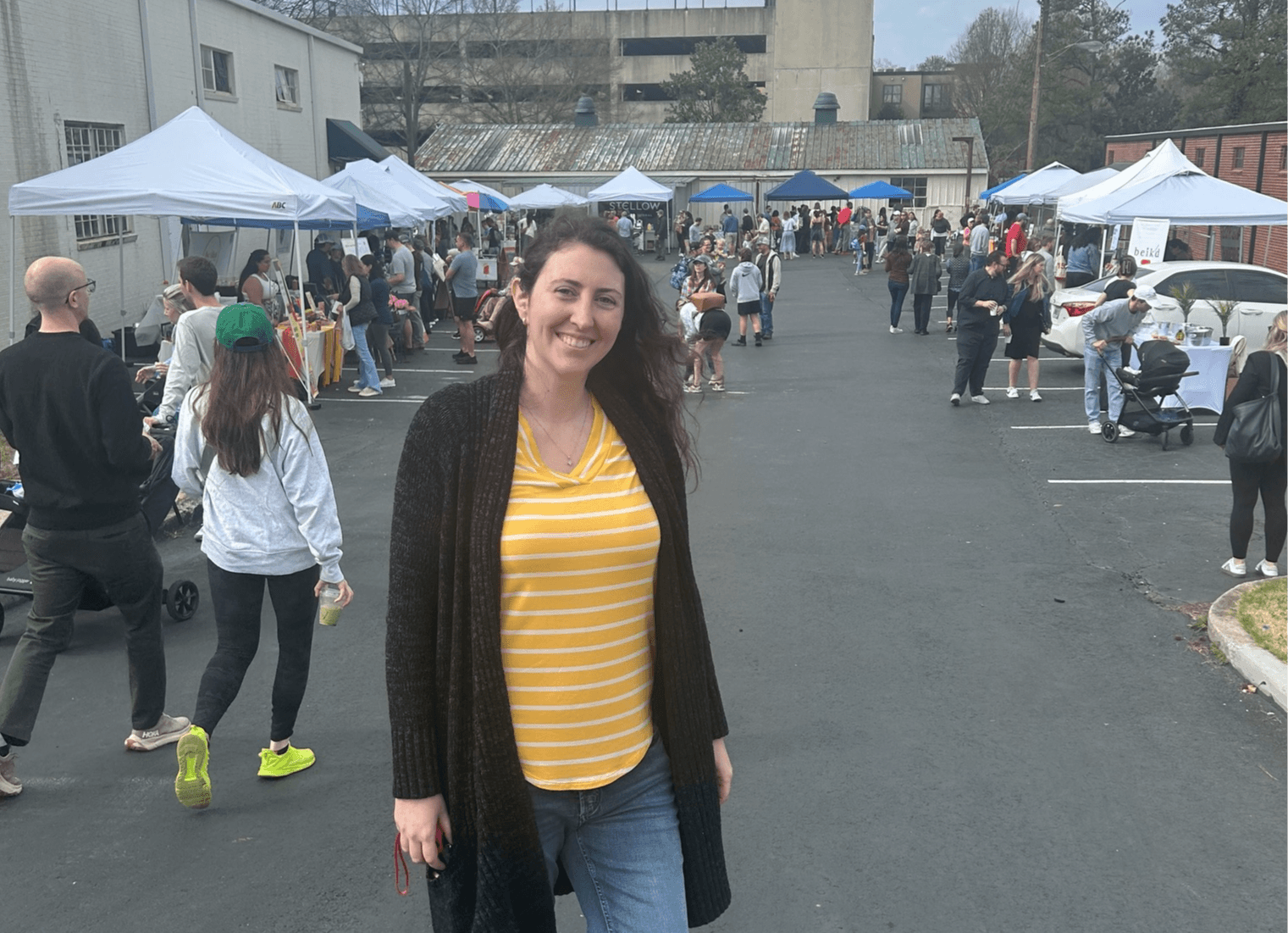 Young woman smiling in front of outdoor market with tents and people, wearing a yellow striped shirt and black coat.