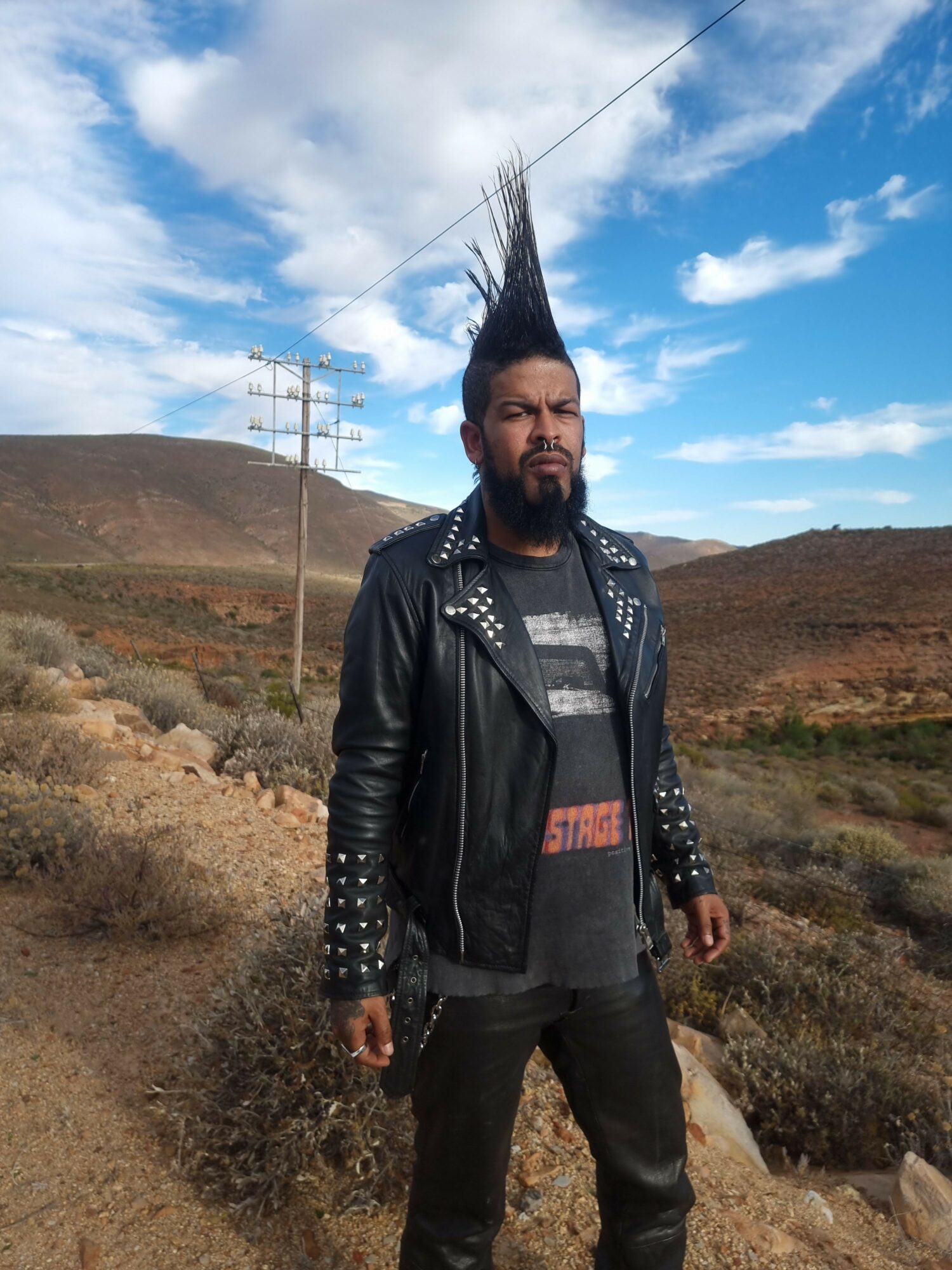 Person with spiked hair and leather jacket standing outdoors with cloudy sky and power lines in background.
