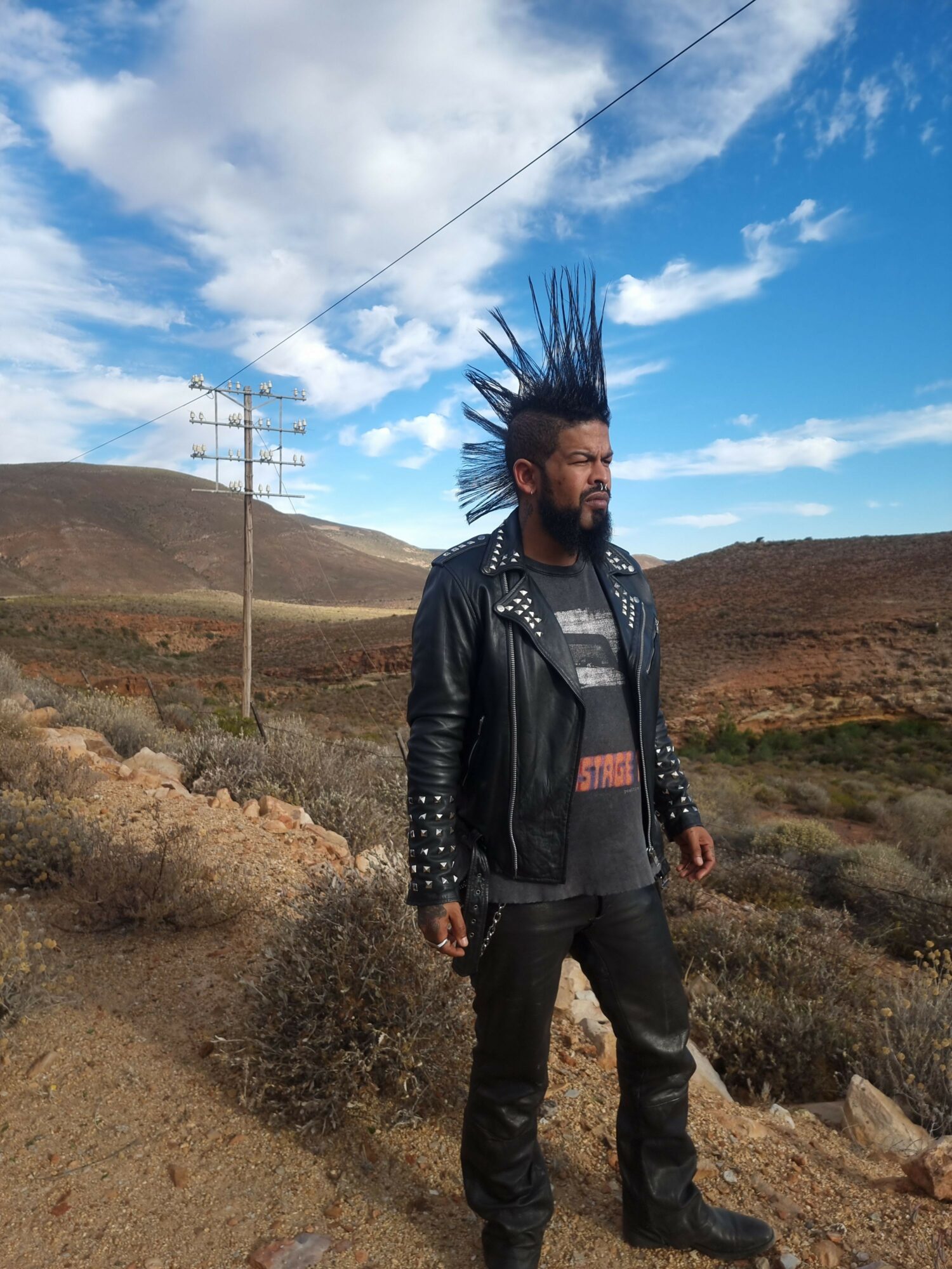 Person with spiked hair, wearing a black leather jacket, standing outdoors on a dirt path with a blue sky and clouds.