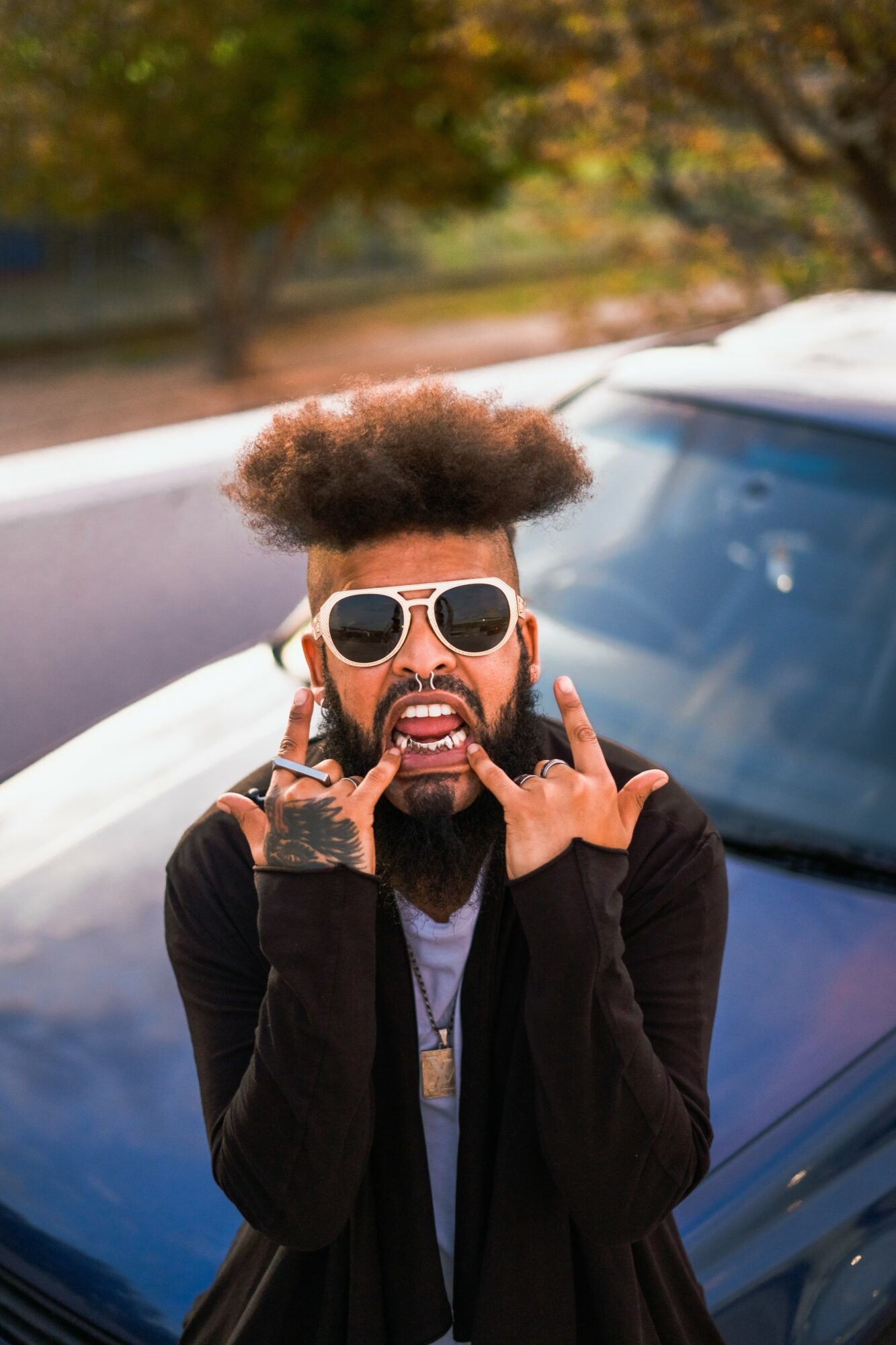 Person with sunglasses and beard making a gesture with hands in front of a car, outdoors with trees in background.