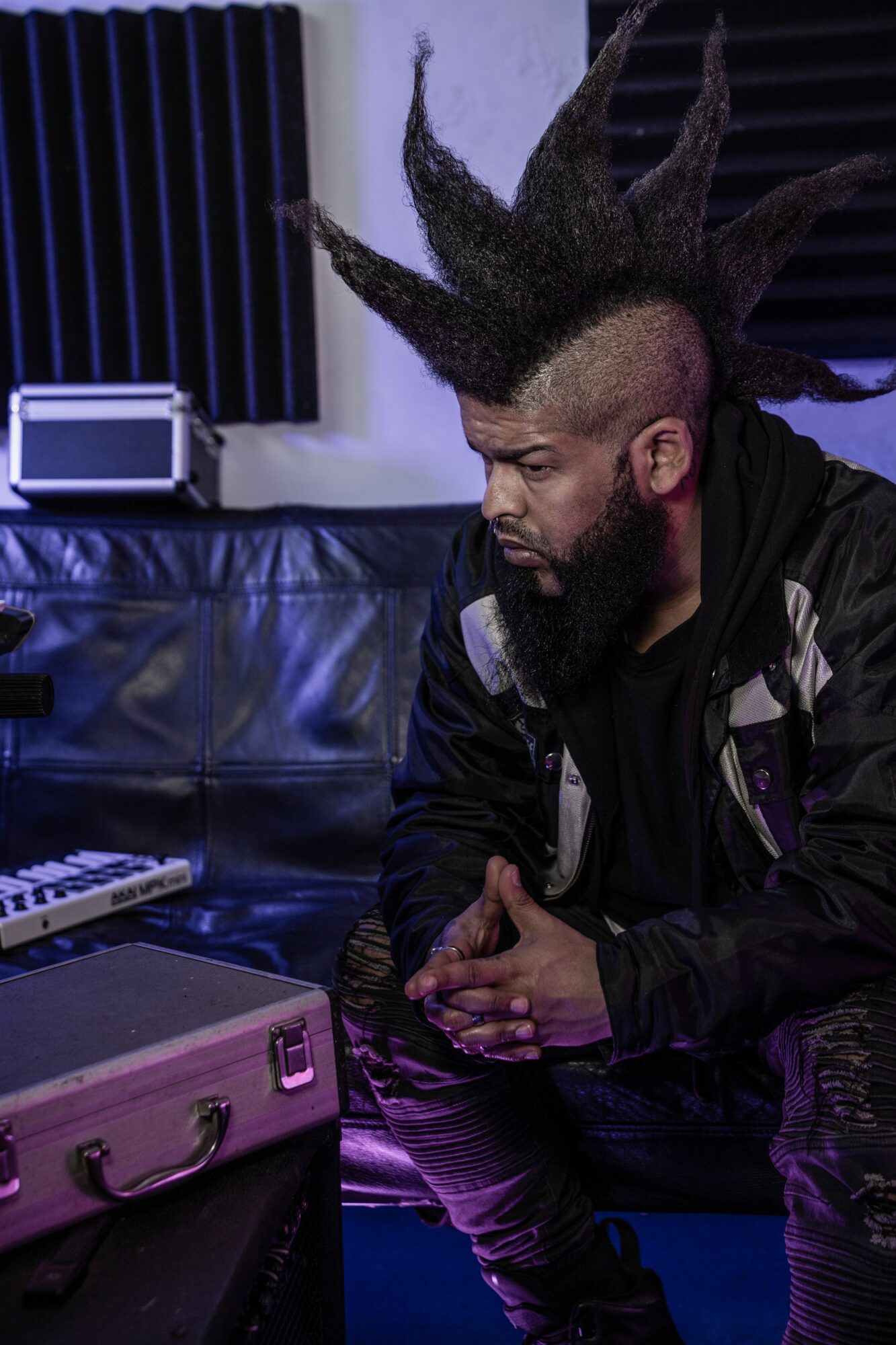 Person with spiked hairstyle sitting on a black leather couch in a music studio, looking down, with equipment around.