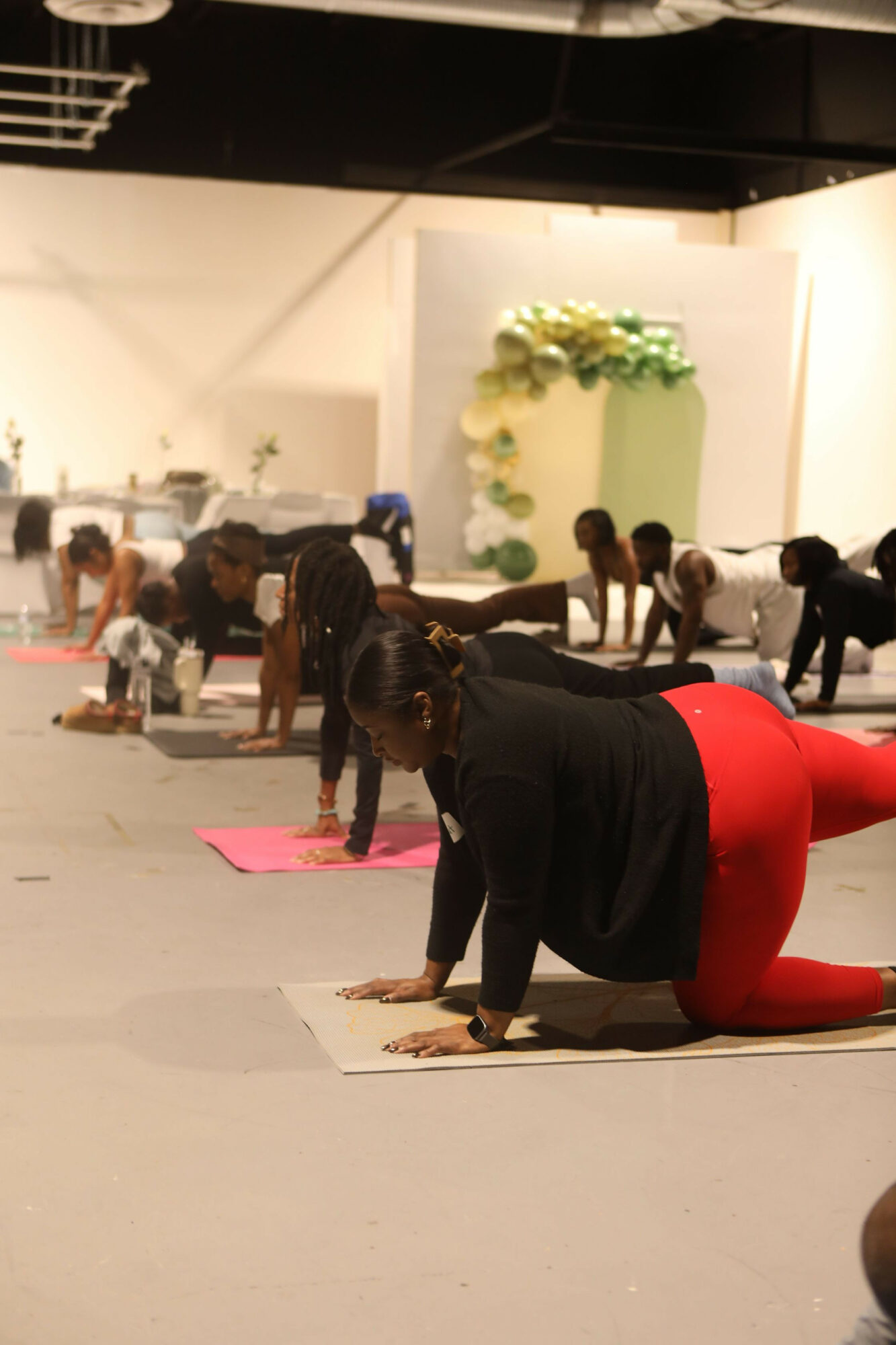 People participating in a yoga class on mats, practicing poses on a spacious indoor floor with a decorative backdrop featuring flowers.