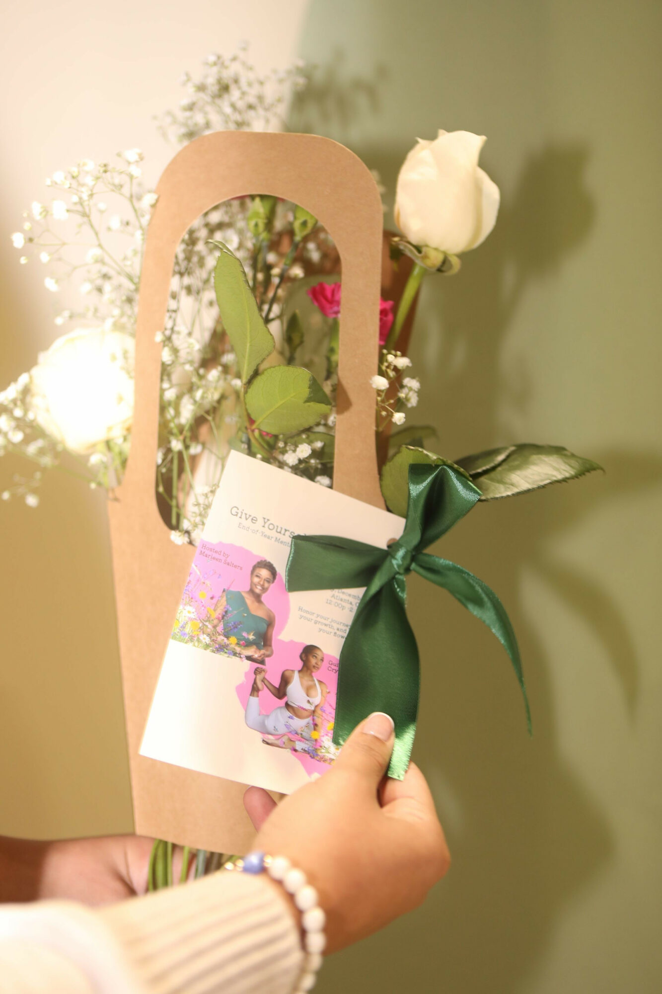 Person holds a gift bag with a white rose and green ribbon, with a card inside, against a light background.