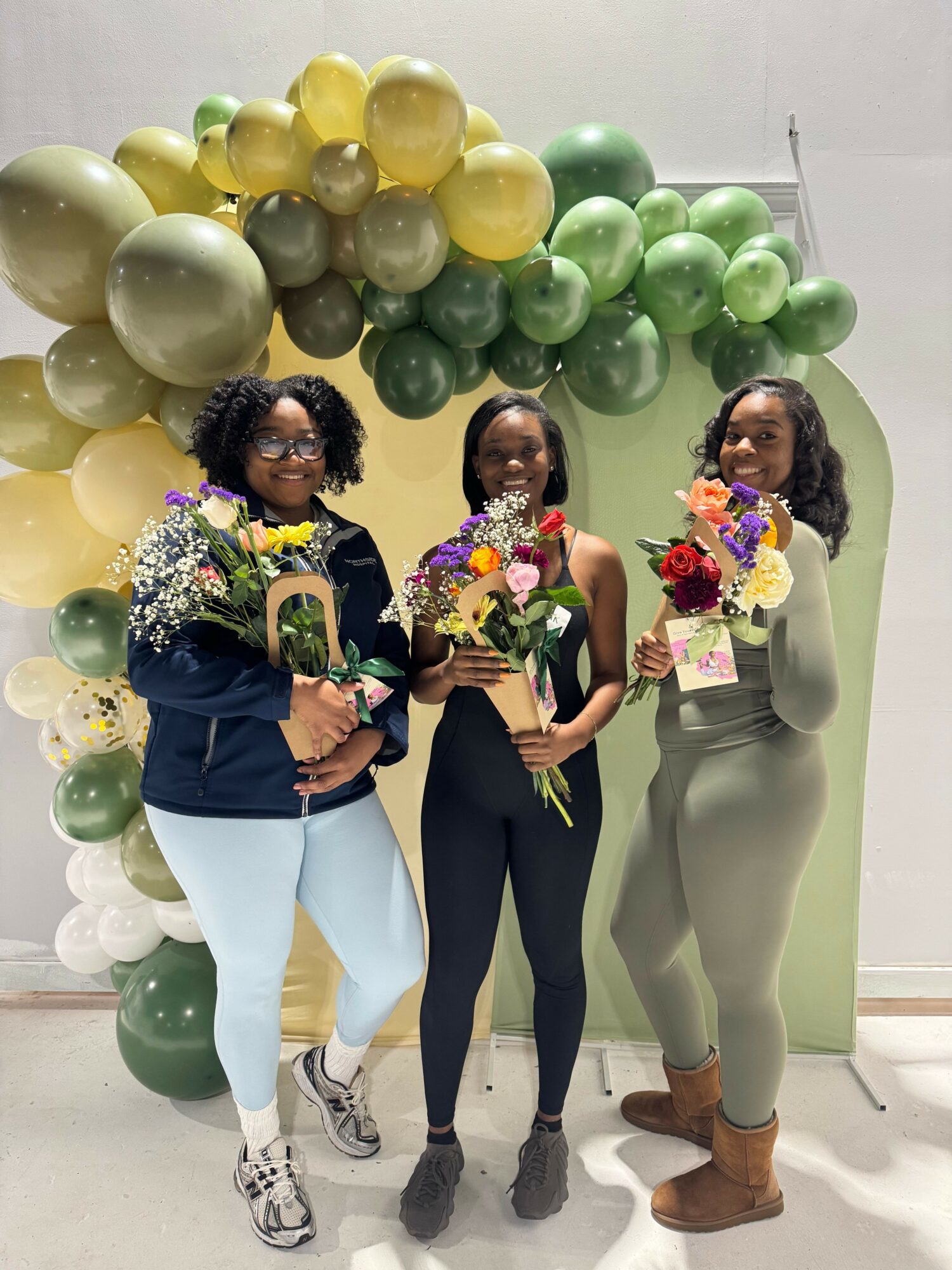 Three women standing in front of green and yellow balloons, holding colorful bouquets, smiling at the camera.
