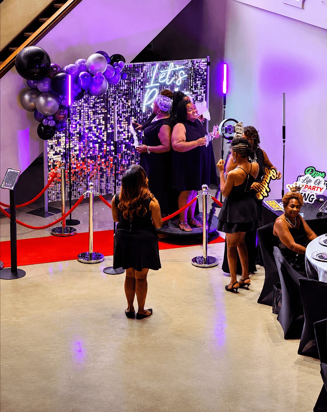 People gather near a decorated backdrop with balloons and a neon sign, celebrating at an indoor event with tables and chairs.
