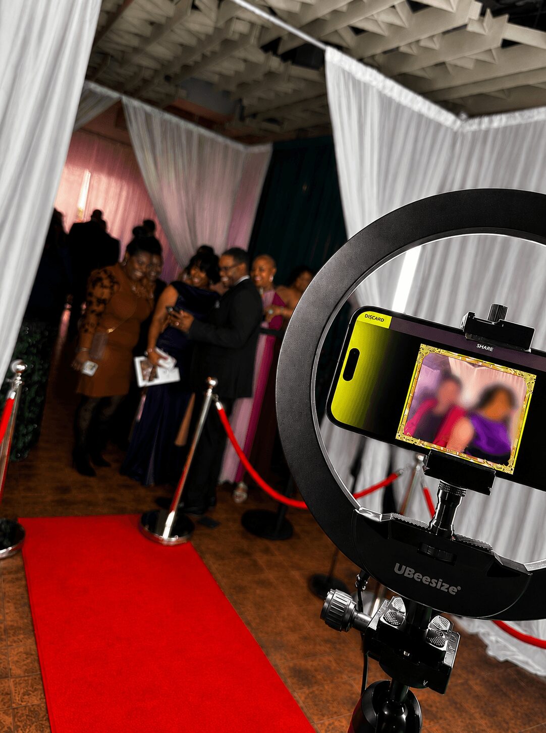 People gather on a red carpet at an indoor event with draped white and pink curtains, and a ring light with a phone in the foreground.