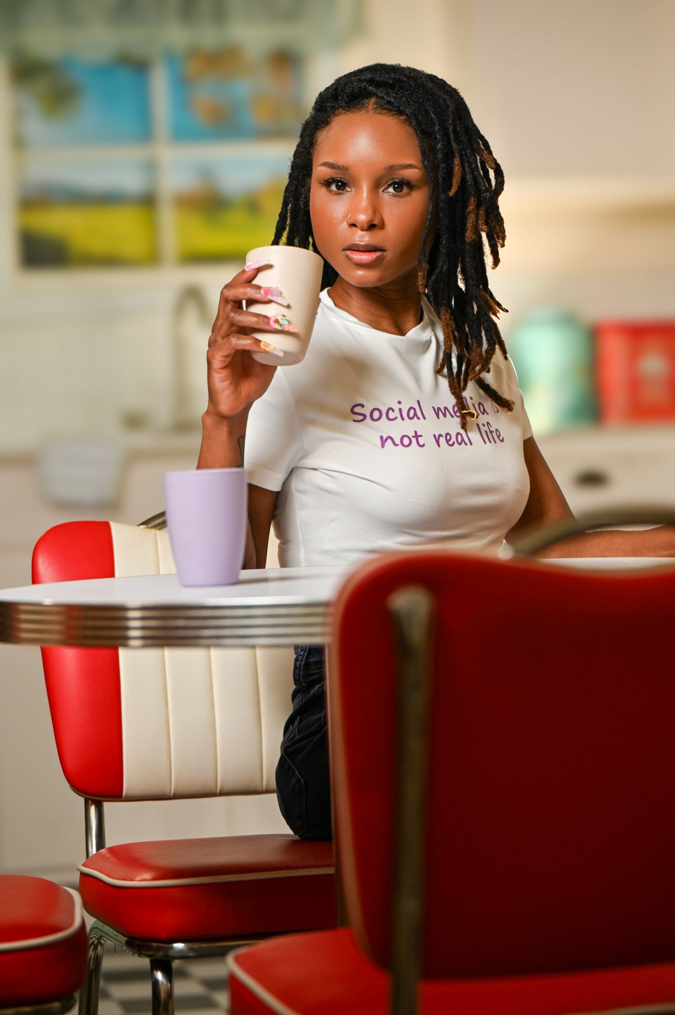 Young woman with dreadlocks holding a white mug, sitting at a table with colorful cups, in a bright room.