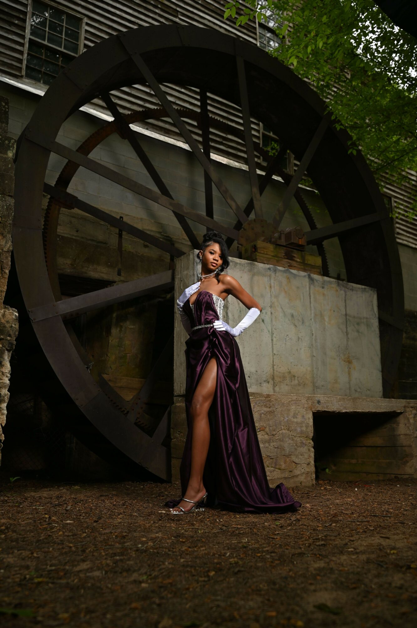 Person in costume standing outdoors near a large water wheel, holding a prop sword, with trees and structures nearby.