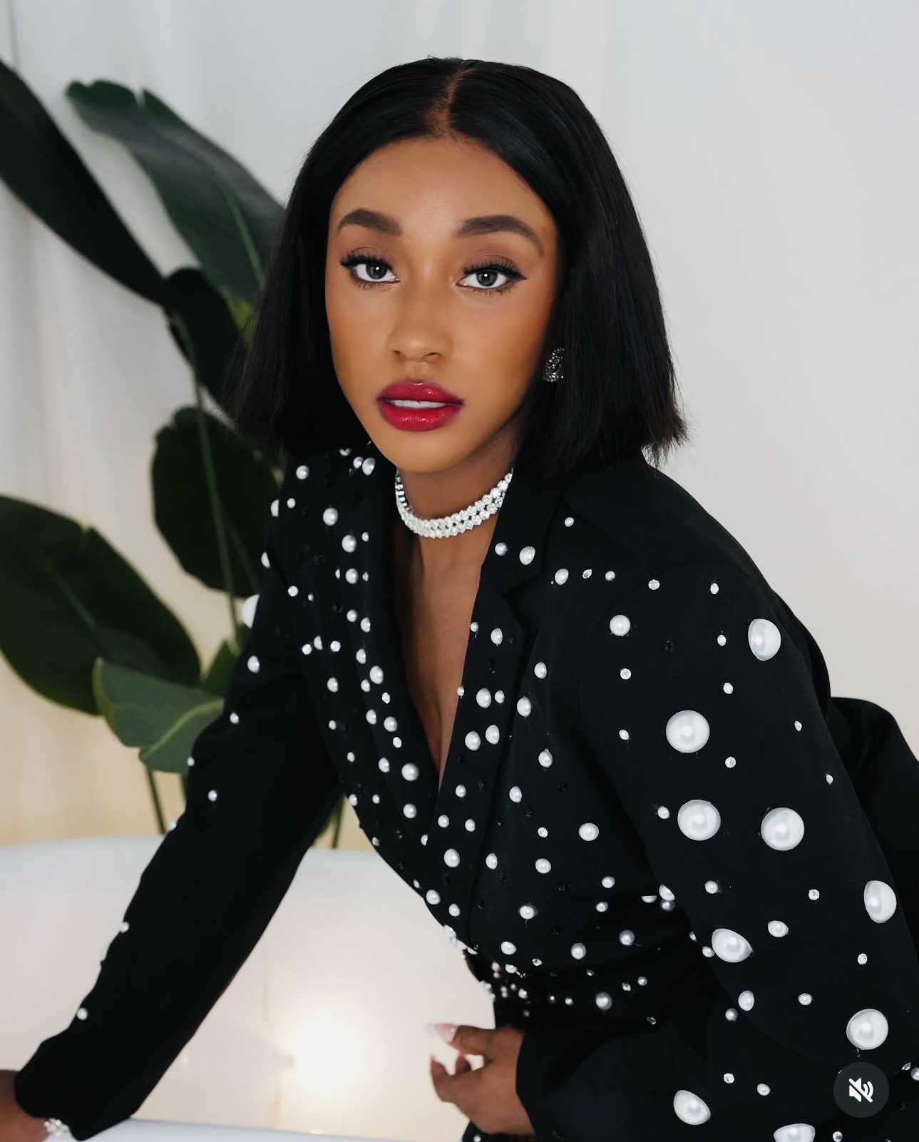 Young woman with black hair and red lipstick, wearing a black polka dot top, posing indoors with a plant in background.