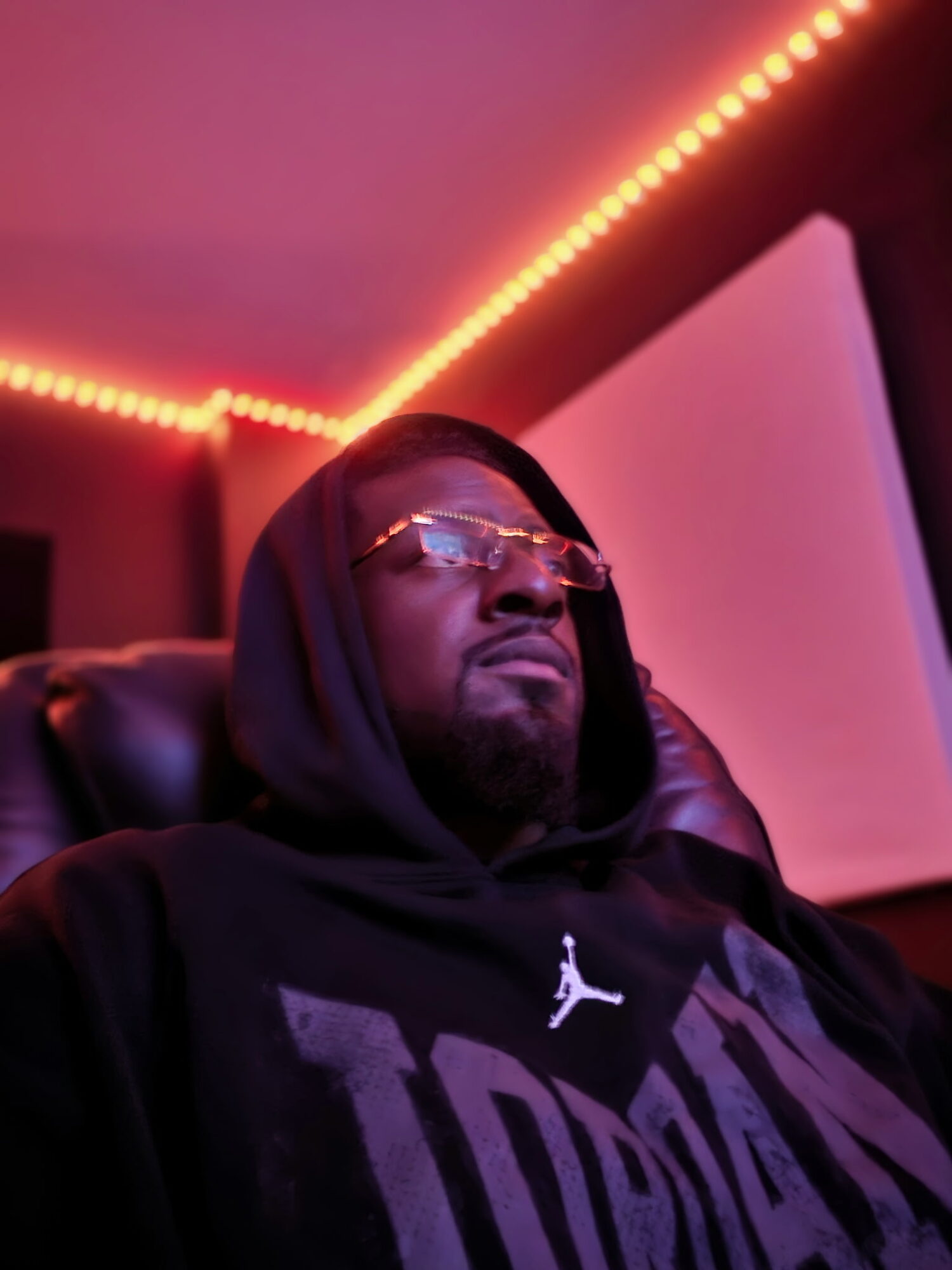 Person wearing glasses and a hoodie, sitting in a room with colorful lighting, looking to the side.