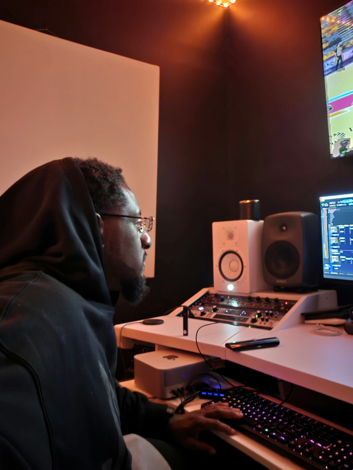 Person with glasses and hoodie working at a computer in a dark room with audio equipment and a screen.