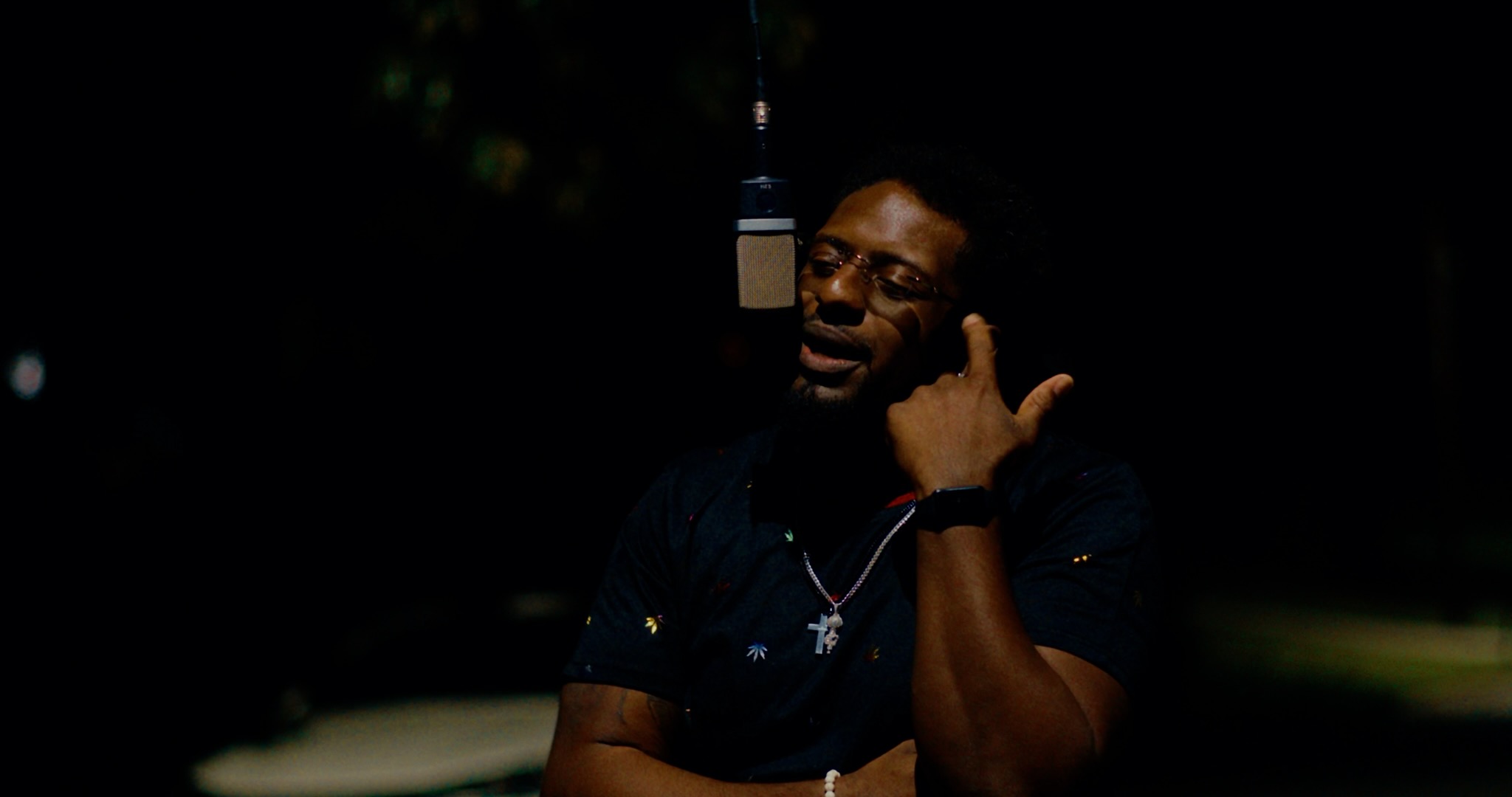 Person with glasses and necklace, talking on a phone in a dark outdoor setting.