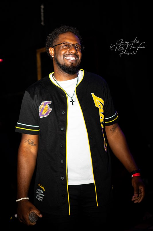 Smiling man with glasses, beard, wearing a black sports jersey with yellow accents, standing in a dark background.