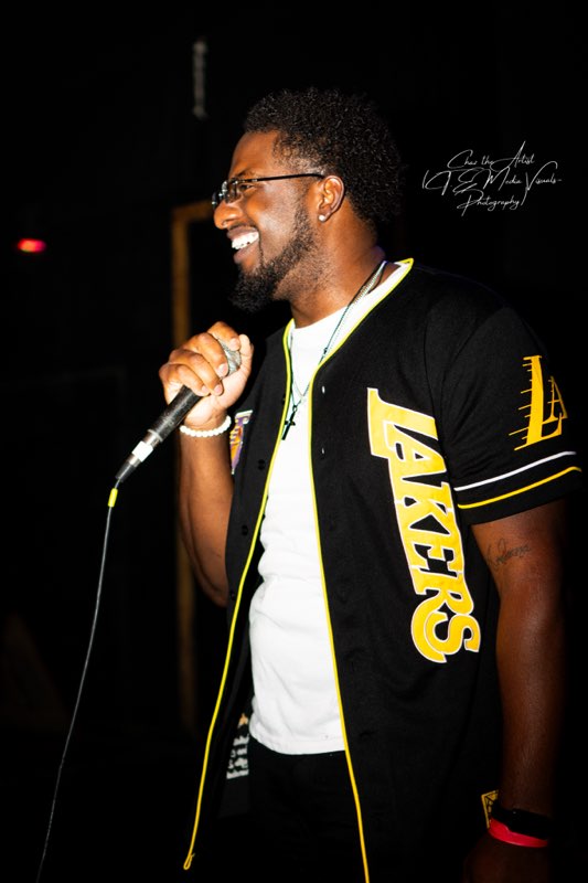 Man with glasses and a beard smiling, holding a microphone, wearing a black jacket with yellow lettering, in a dark setting.
