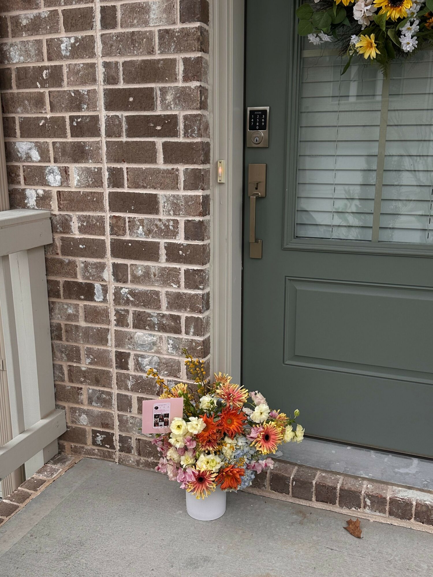 Flower arrangement with colorful flowers on a white container outside a green door, brick wall background.