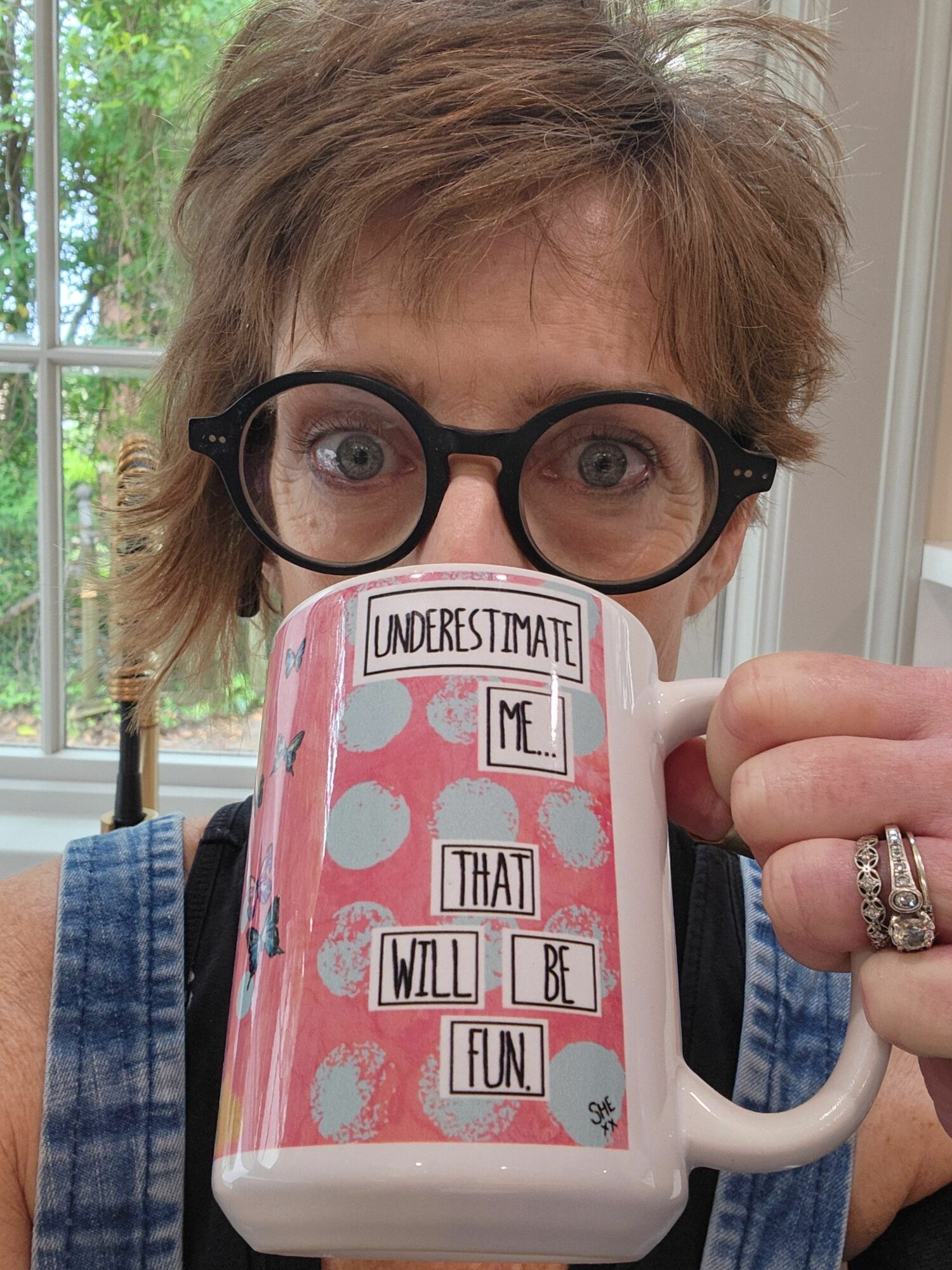 Person with glasses holding a pink mug with stickers, standing near a window with trees outside.