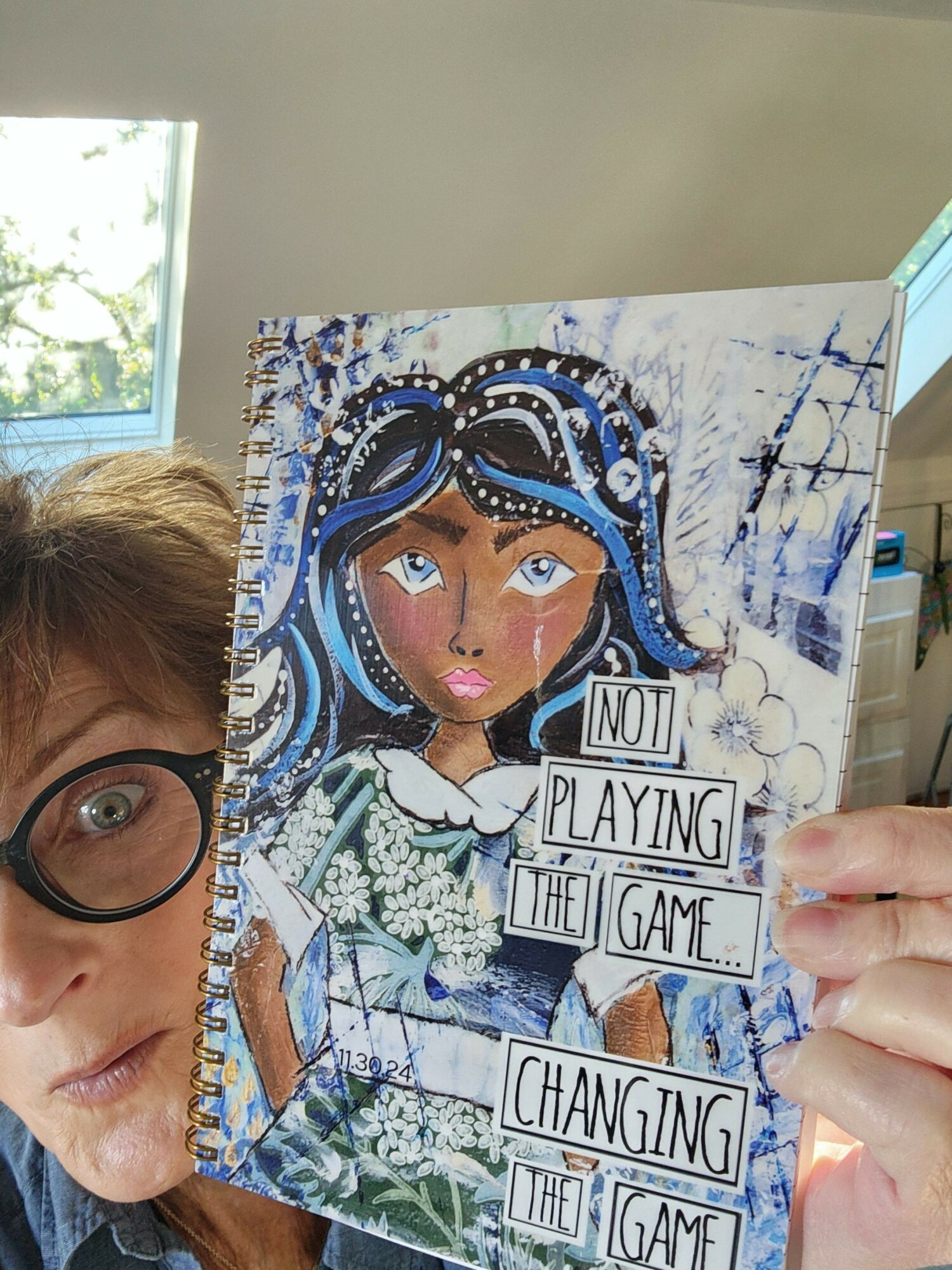 Person with glasses holding a colorful illustrated book, partially visible face, background with window and trees.
