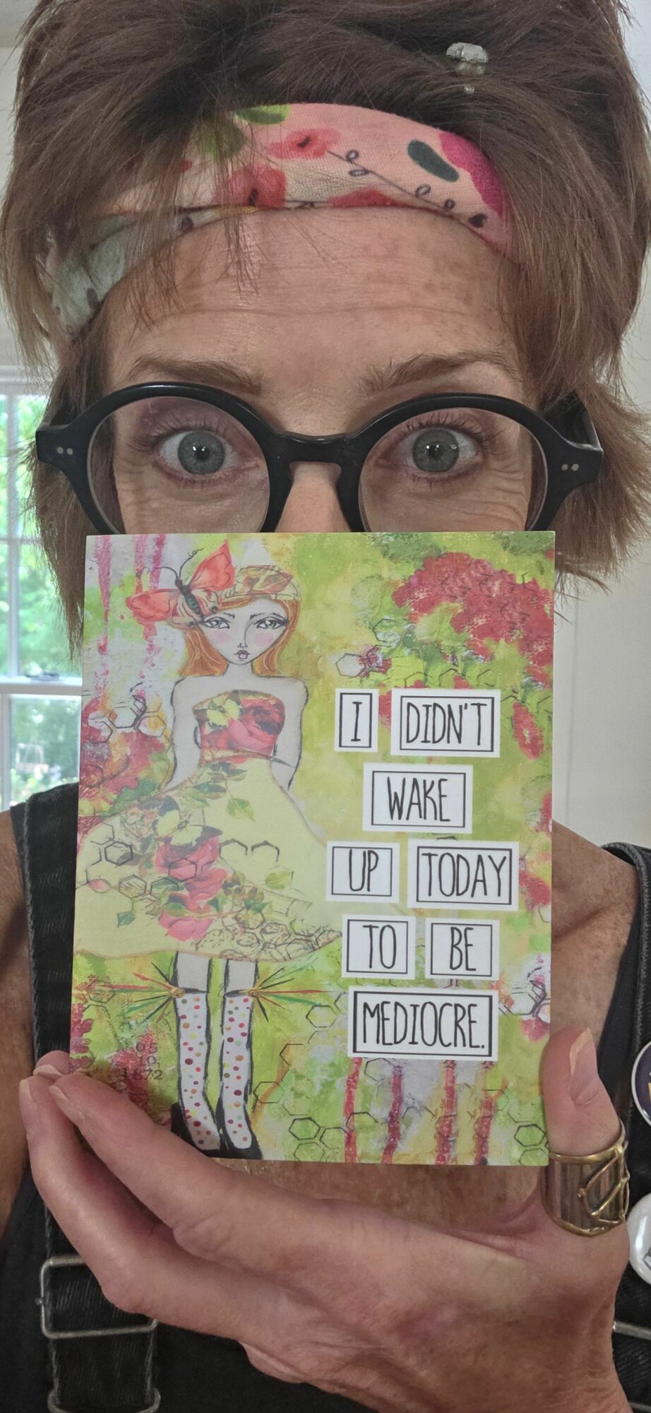 Person with glasses holding a colorful card with text, face partially covered by the card, background shows a window.