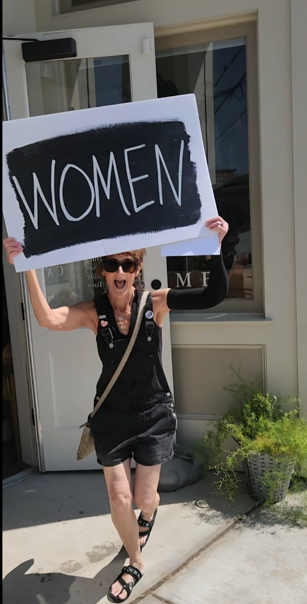 Person holding a sign that says WOMEN, standing outside near a window and plants.