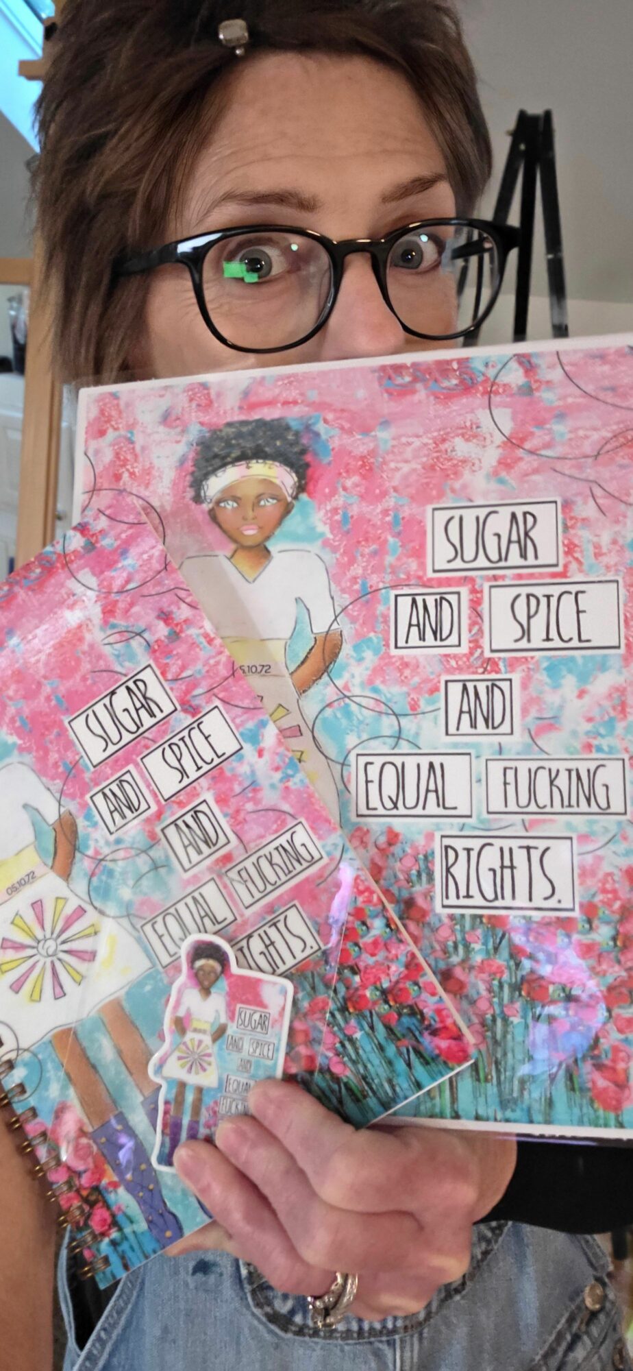 Woman with glasses holding colorful scrapbook pages with text and images, background shows a room with shelves.