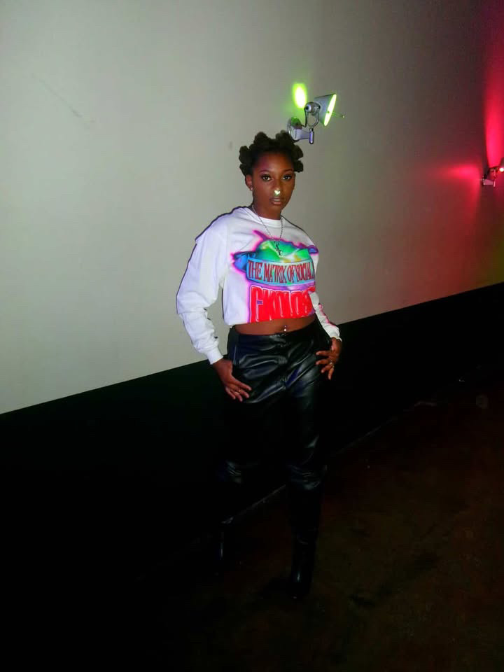 Woman standing against a plain wall with colorful lights, wearing a white sweatshirt and black pants.