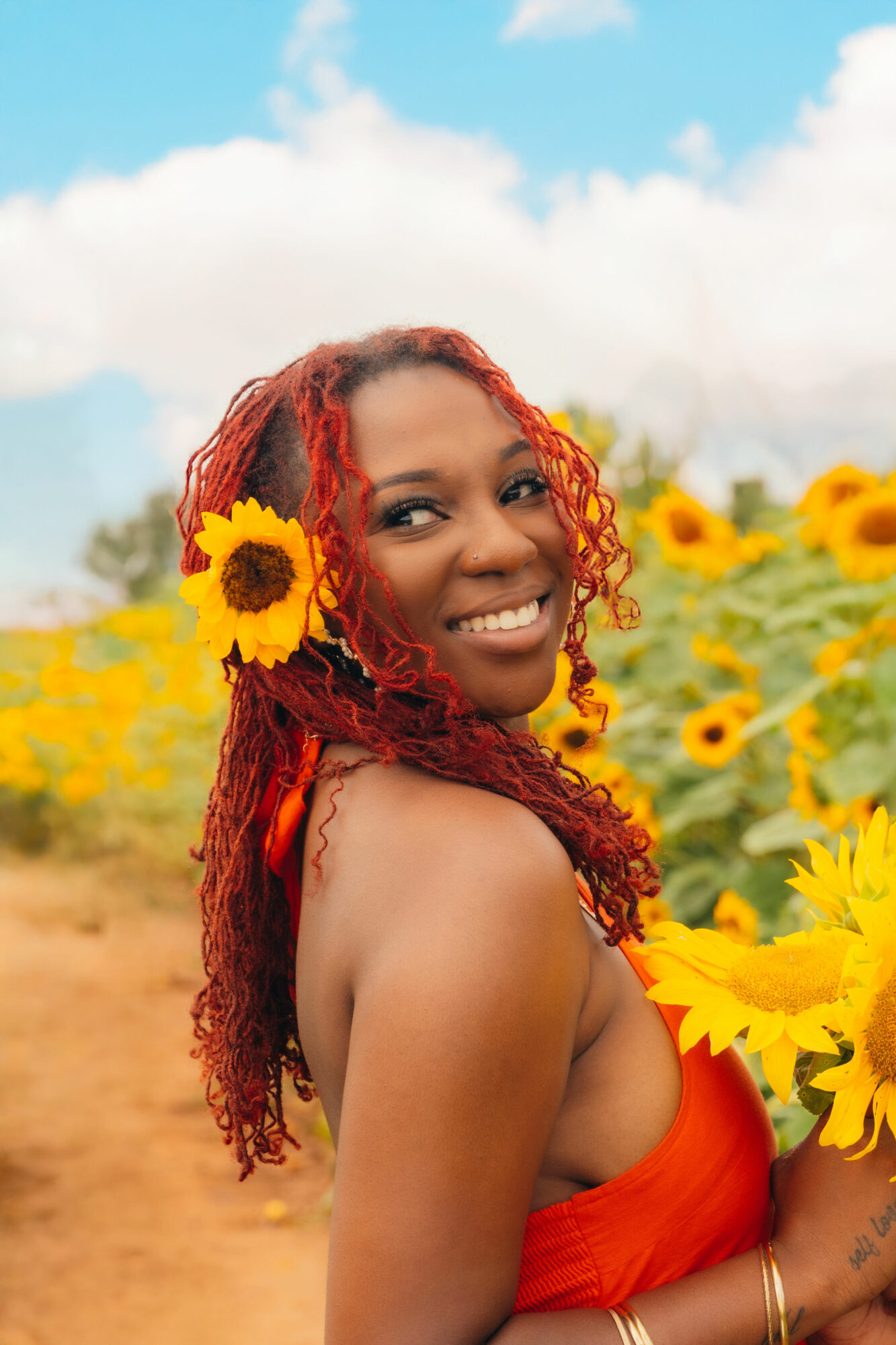 Smiling woman with red curly hair in a sunflower field, holding sunflowers, with a blue sky and clouds behind her.