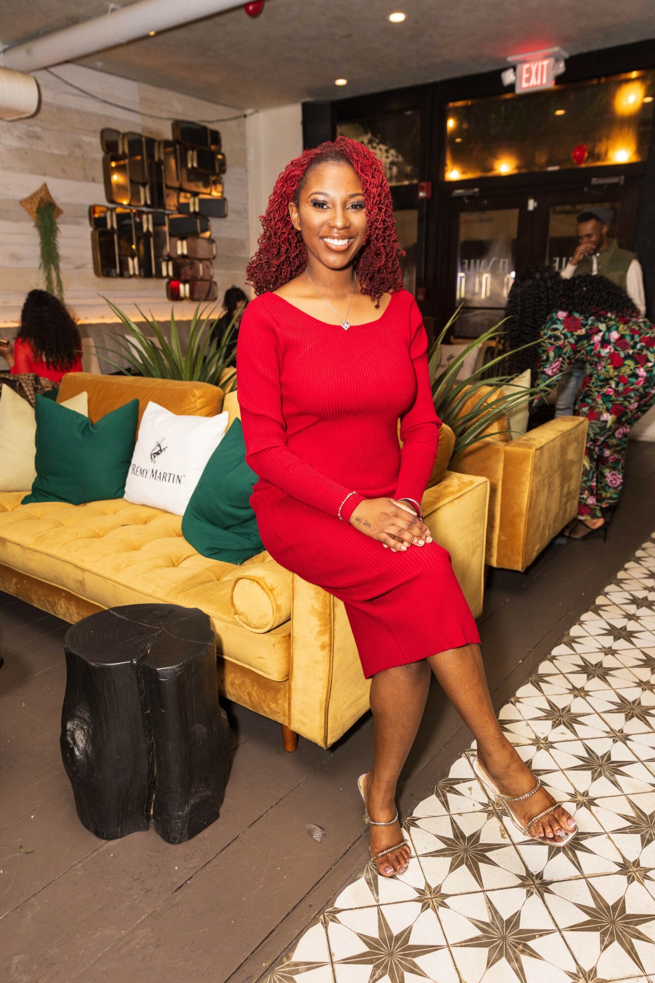 Woman with curly red hair in red dress sitting on a yellow sofa in a decorated indoor space.