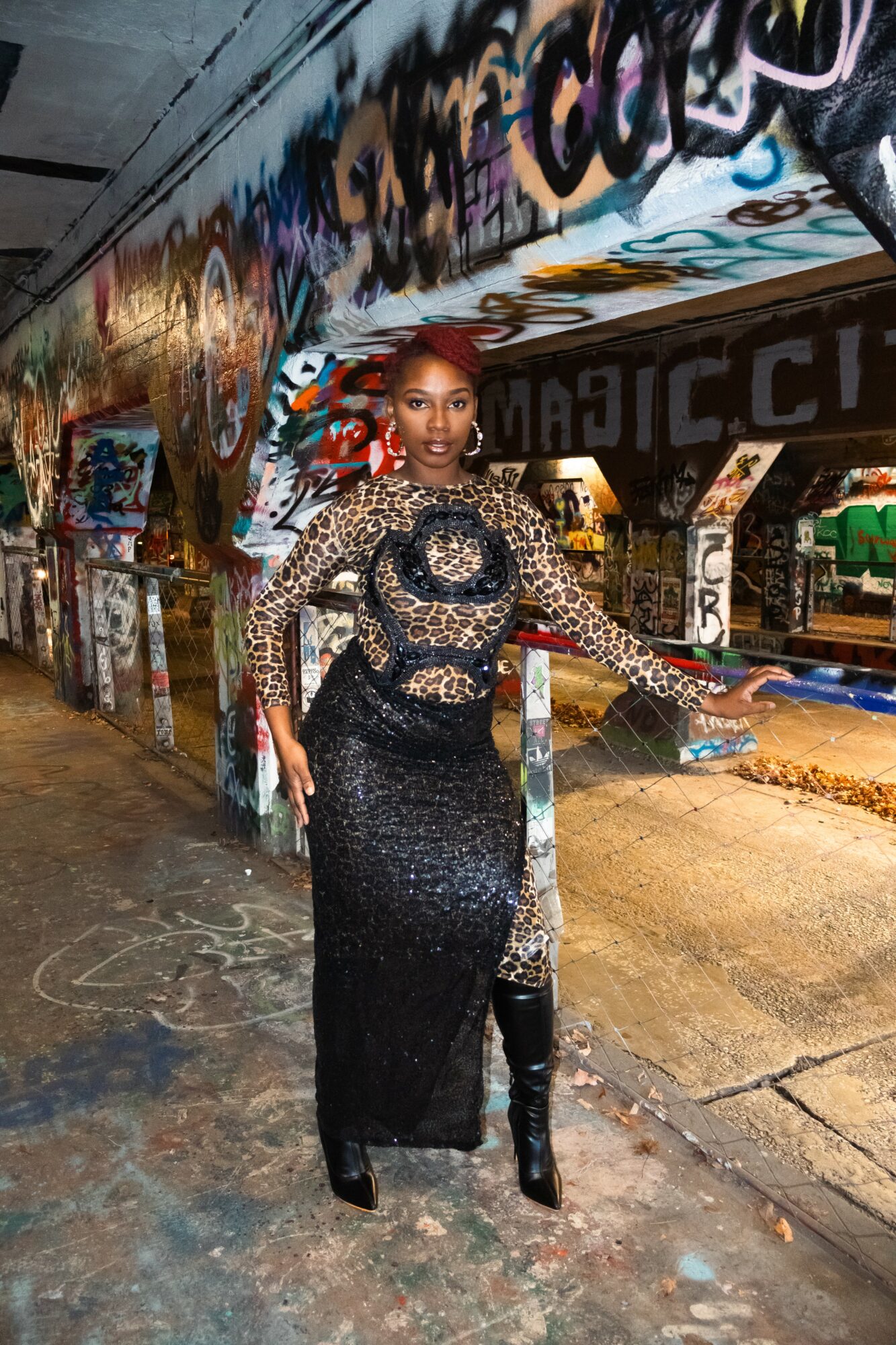 Woman in a black and gold dress with a sheer, patterned overlay stands in a graffiti-covered tunnel, holding a clutch bag.