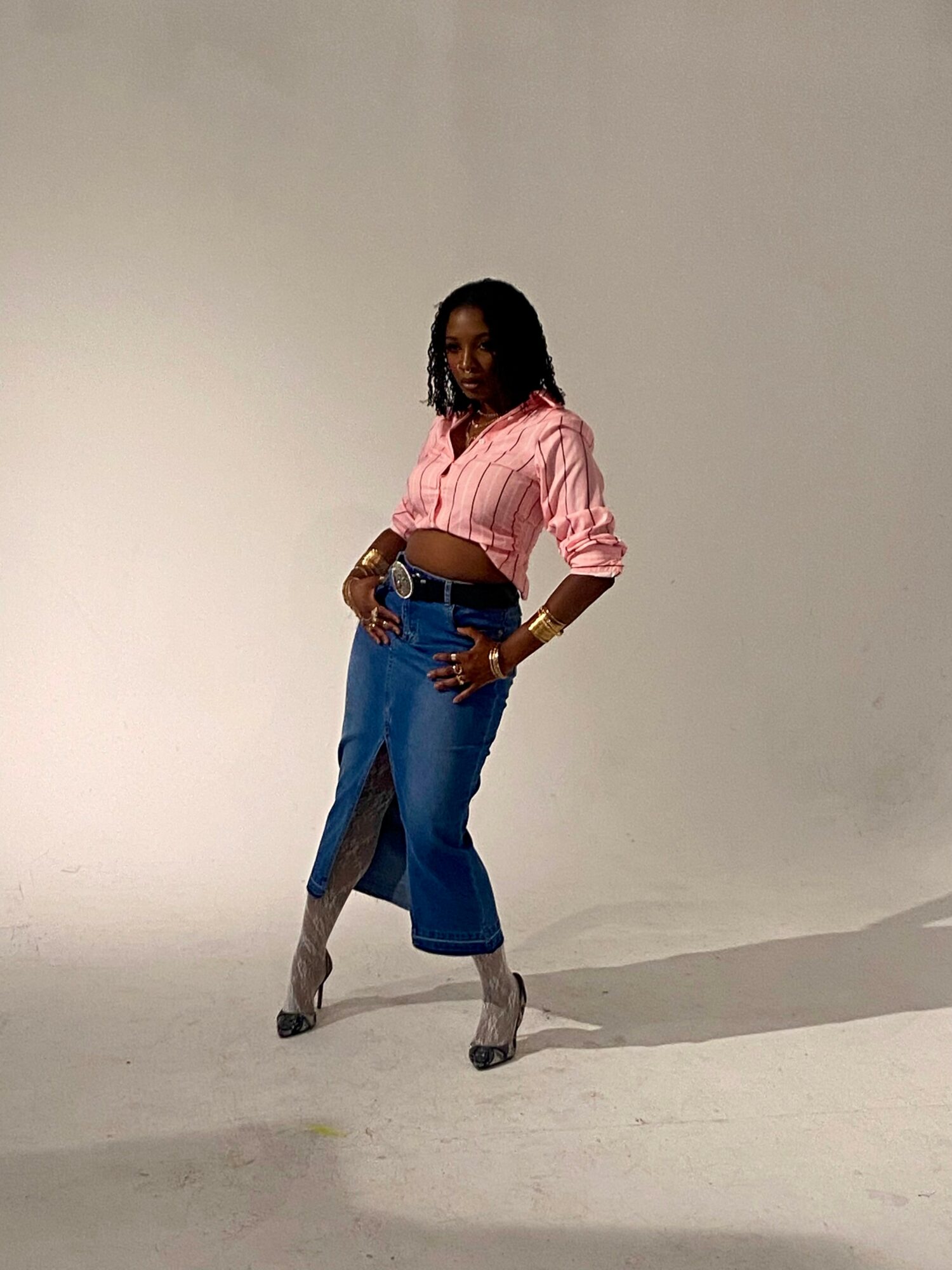 Woman in pink jacket and blue jeans standing with hands on hips against a plain background.