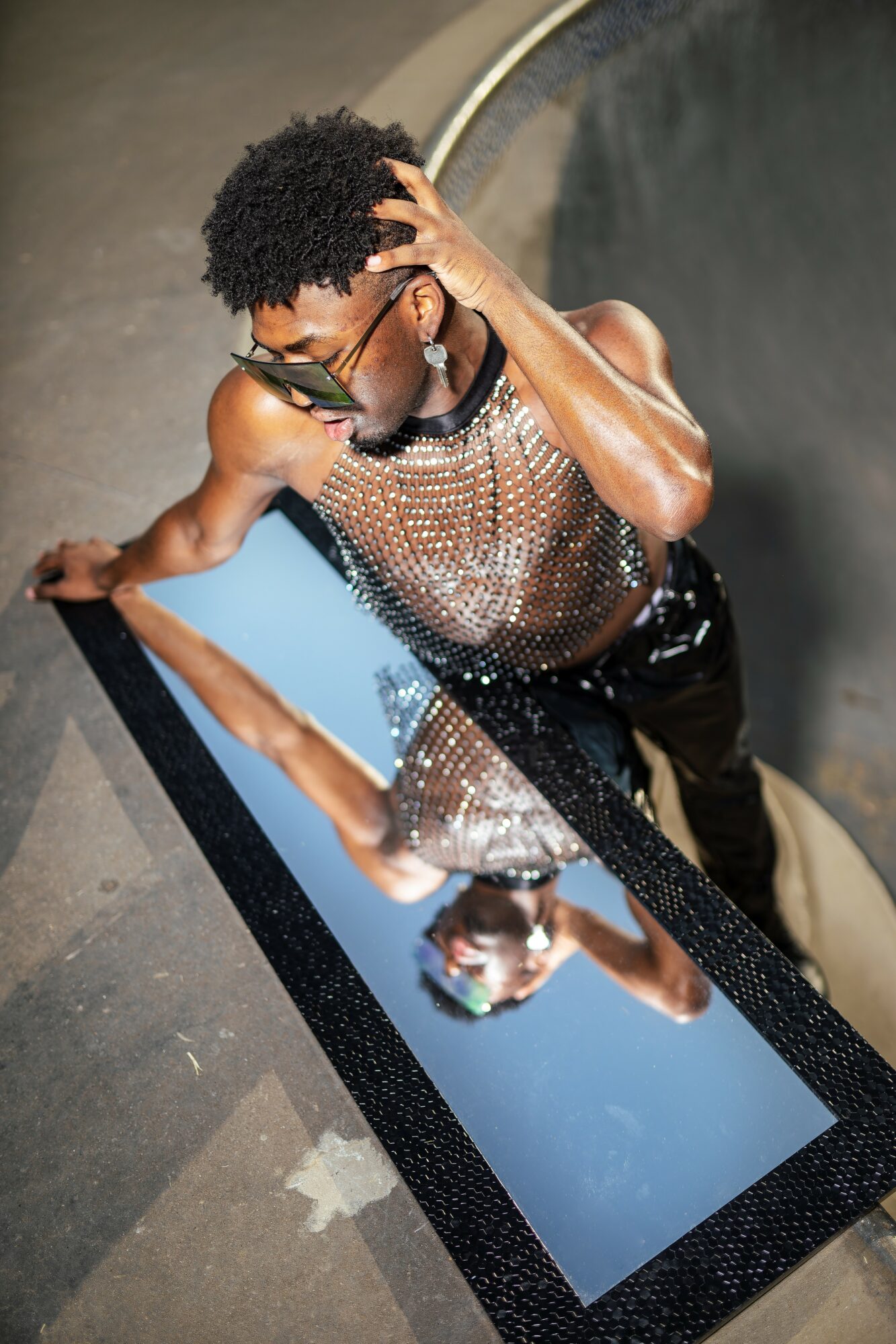Person with curly hair and glasses looks into a mirror, touching their head, wearing a shiny top, reflection visible.