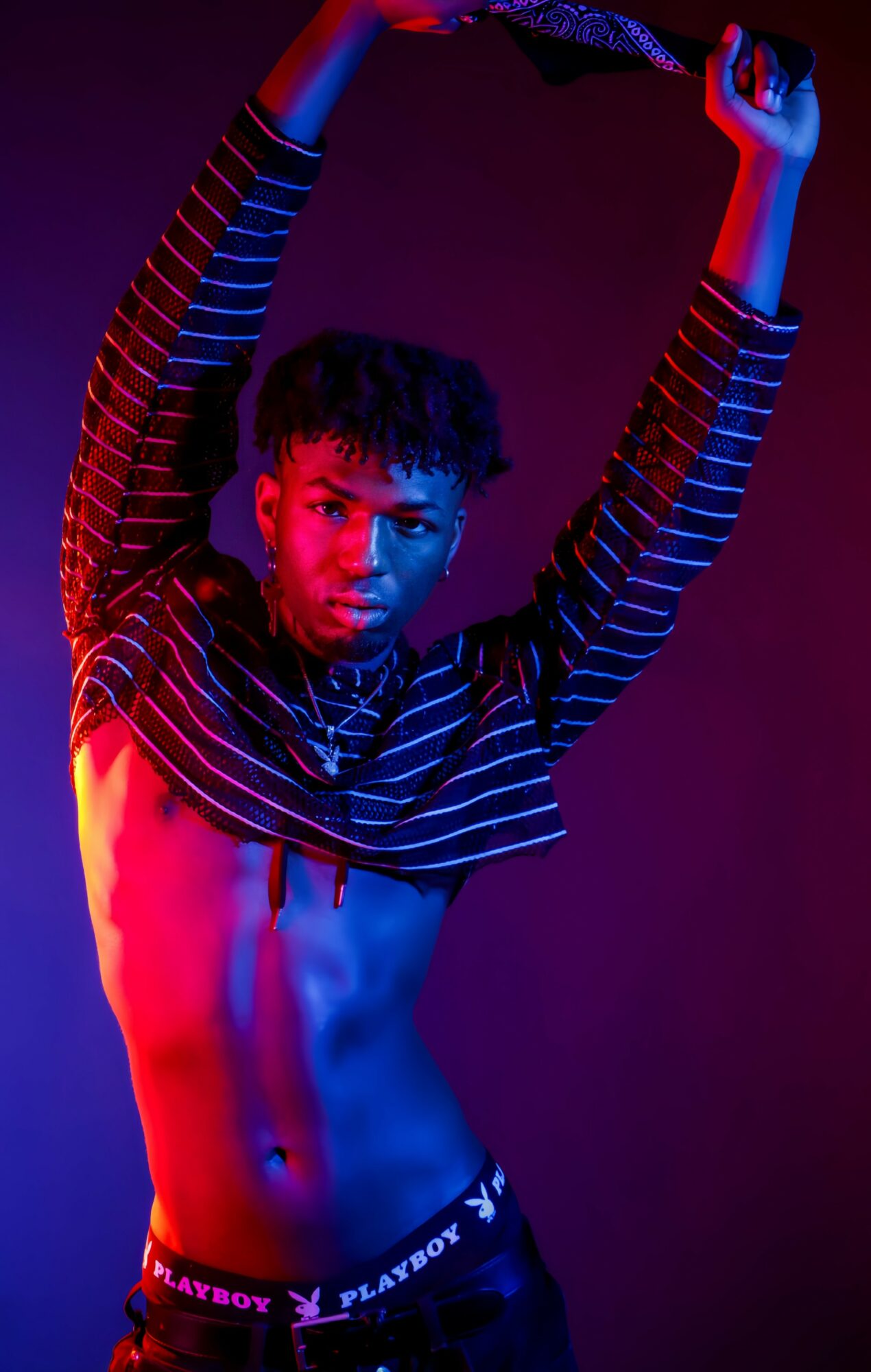 Person with arms raised, wearing striped shirt, illuminated by colorful lighting, exposing midriff, against dark background.