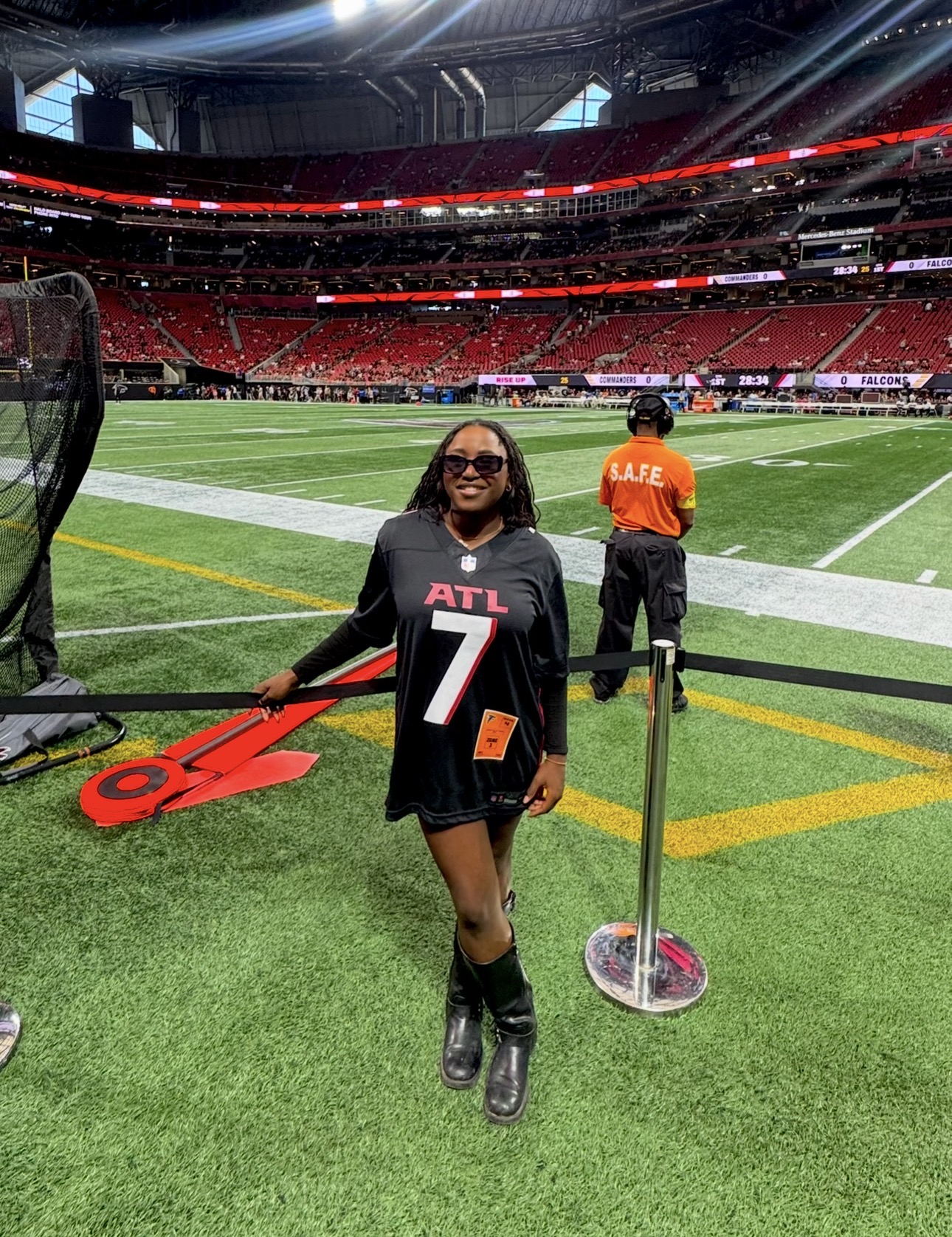 Young girl in black Atlanta Falcons jersey and sunglasses standing on football field with stadium seating in background.