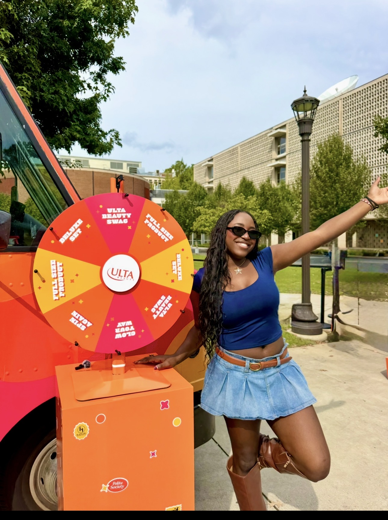 Woman with long dark hair, sunglasses, blue top, and denim skirt, smiling outdoors with a wheel and orange box, arm raised.