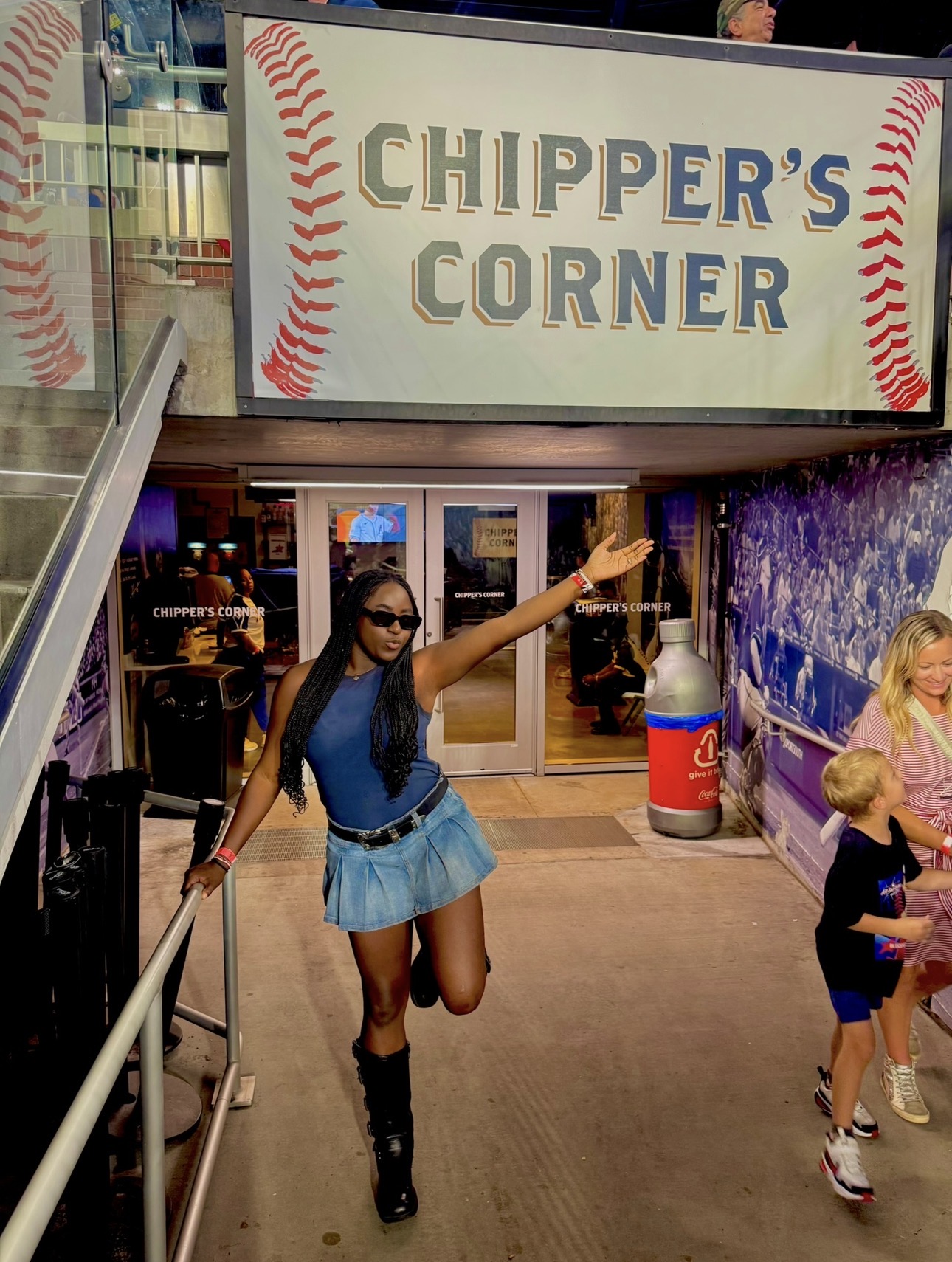 Woman in sunglasses and blue outfit with a skirt, raising her arm inside a baseball-themed area, with children nearby.