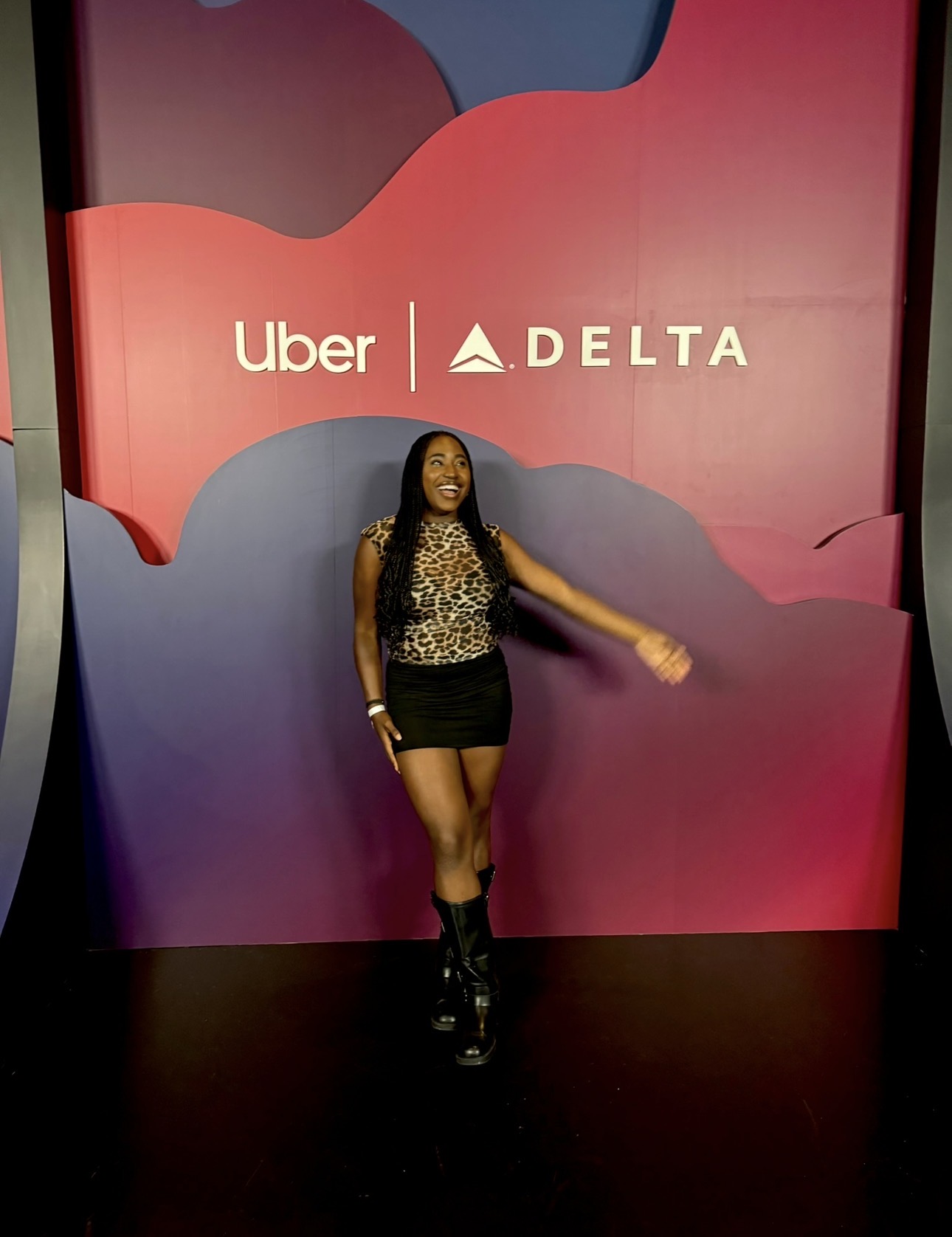 Woman standing in front of a colorful backdrop with Uber and Delta logos, smiling, wearing a patterned top and black skirt.