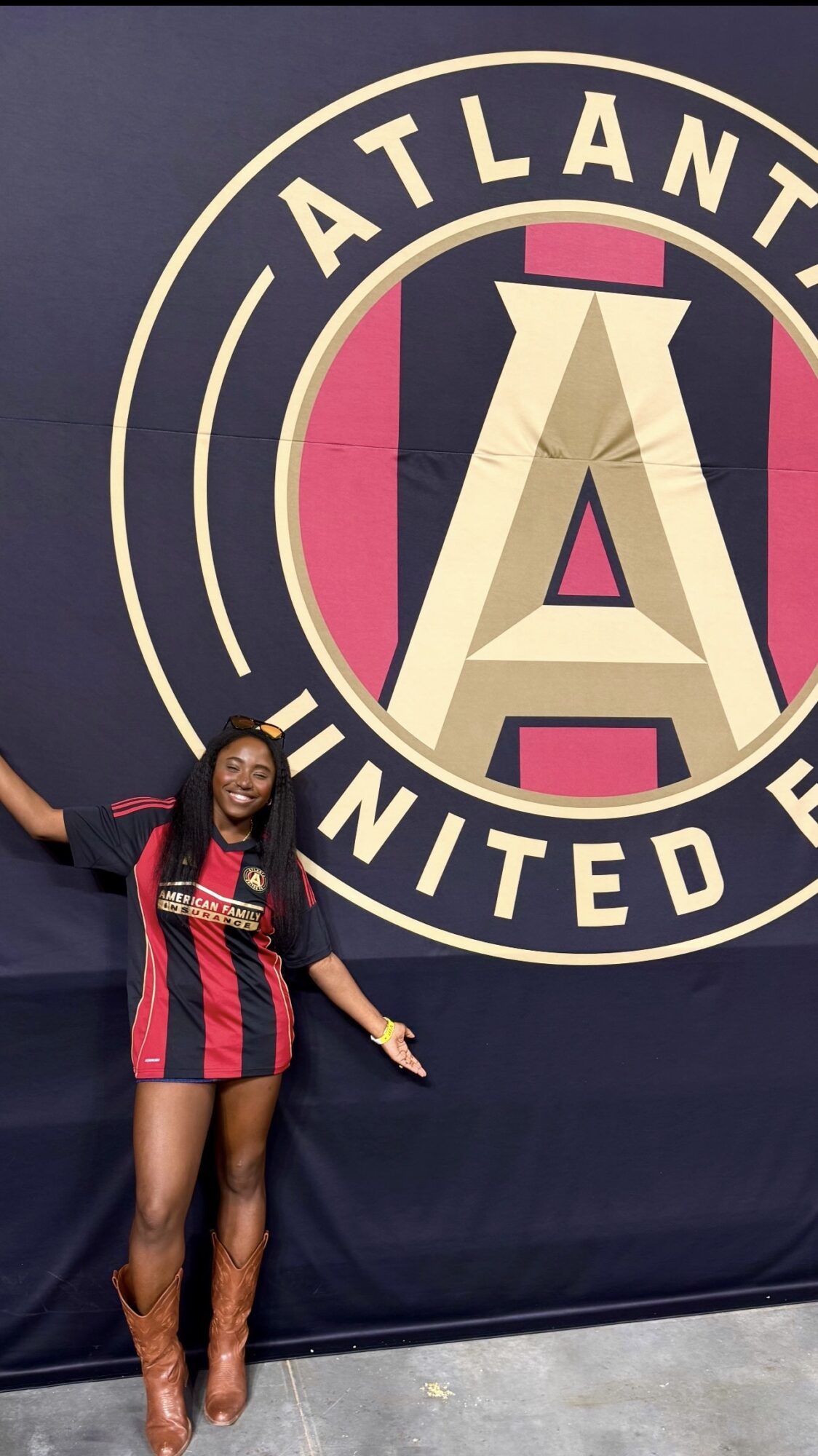 Woman standing in front of Atlanta United FC logo, smiling, wearing a red and blue striped jersey, brown boots, arms outstretched.
