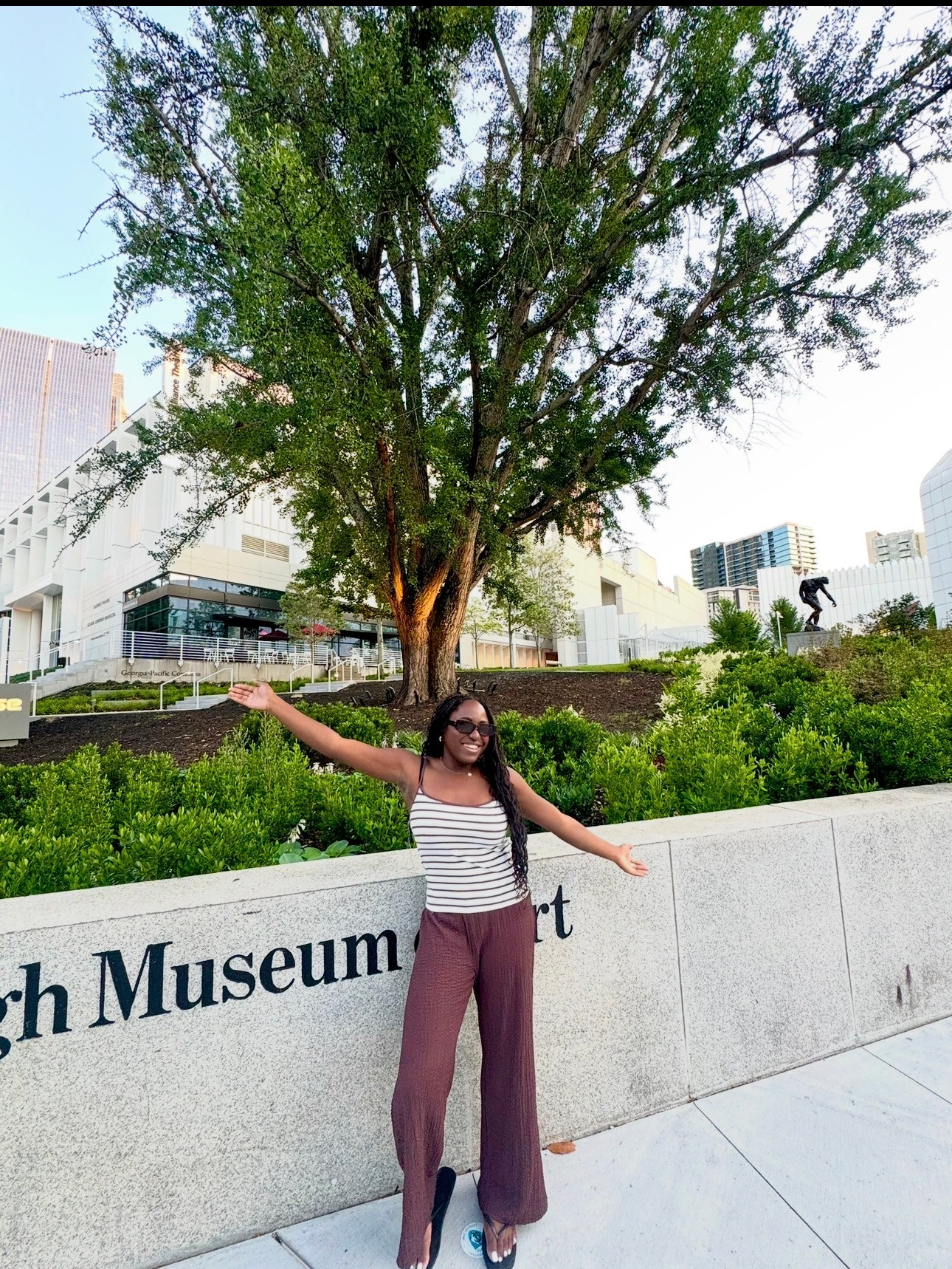 Person standing in front of a large tree and a museum, smiling with arms outstretched, wearing glasses, striped top, and wide pants.