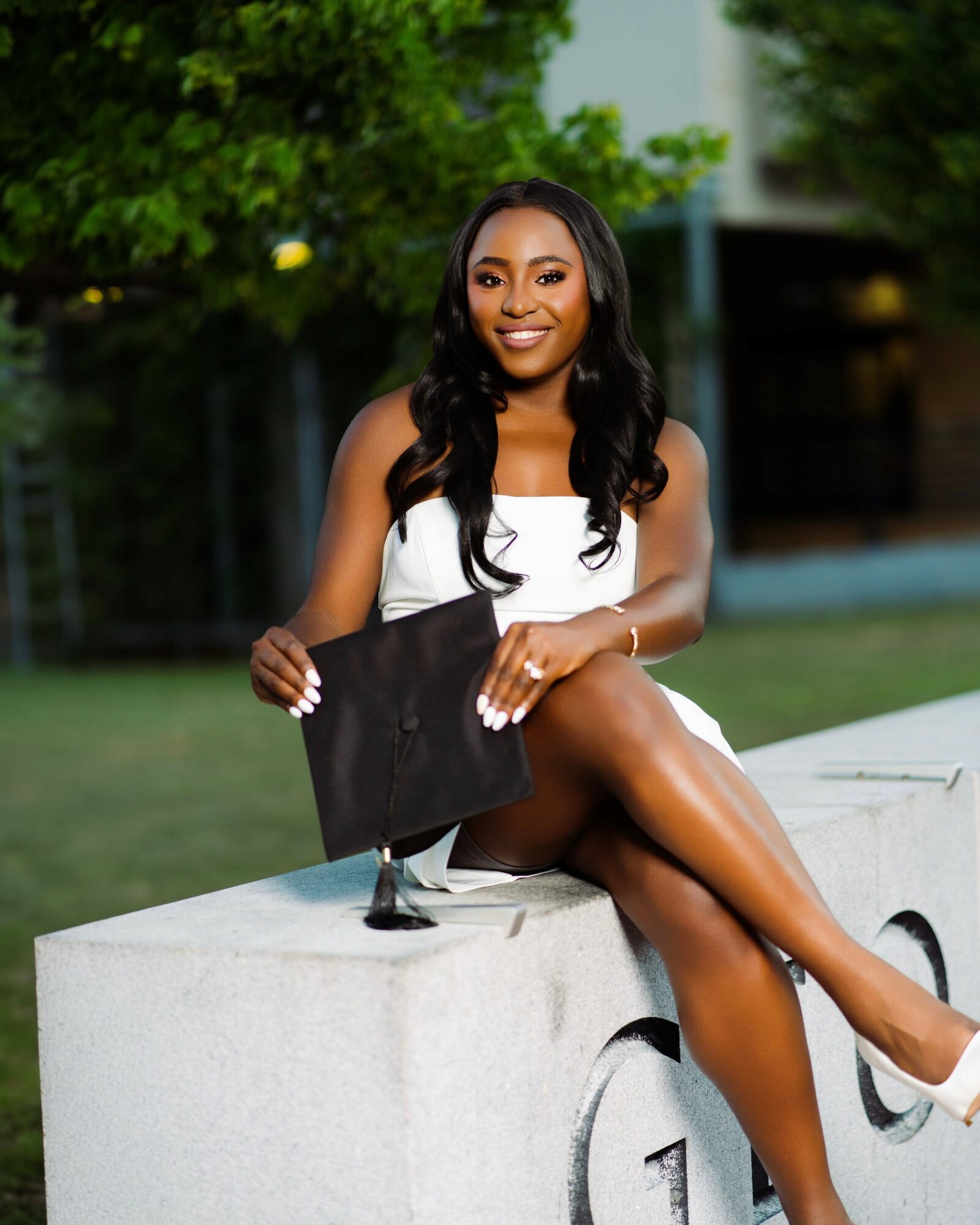 Young woman sitting on a concrete ledge outdoors, smiling, holding a black folder, with trees and a building in background.