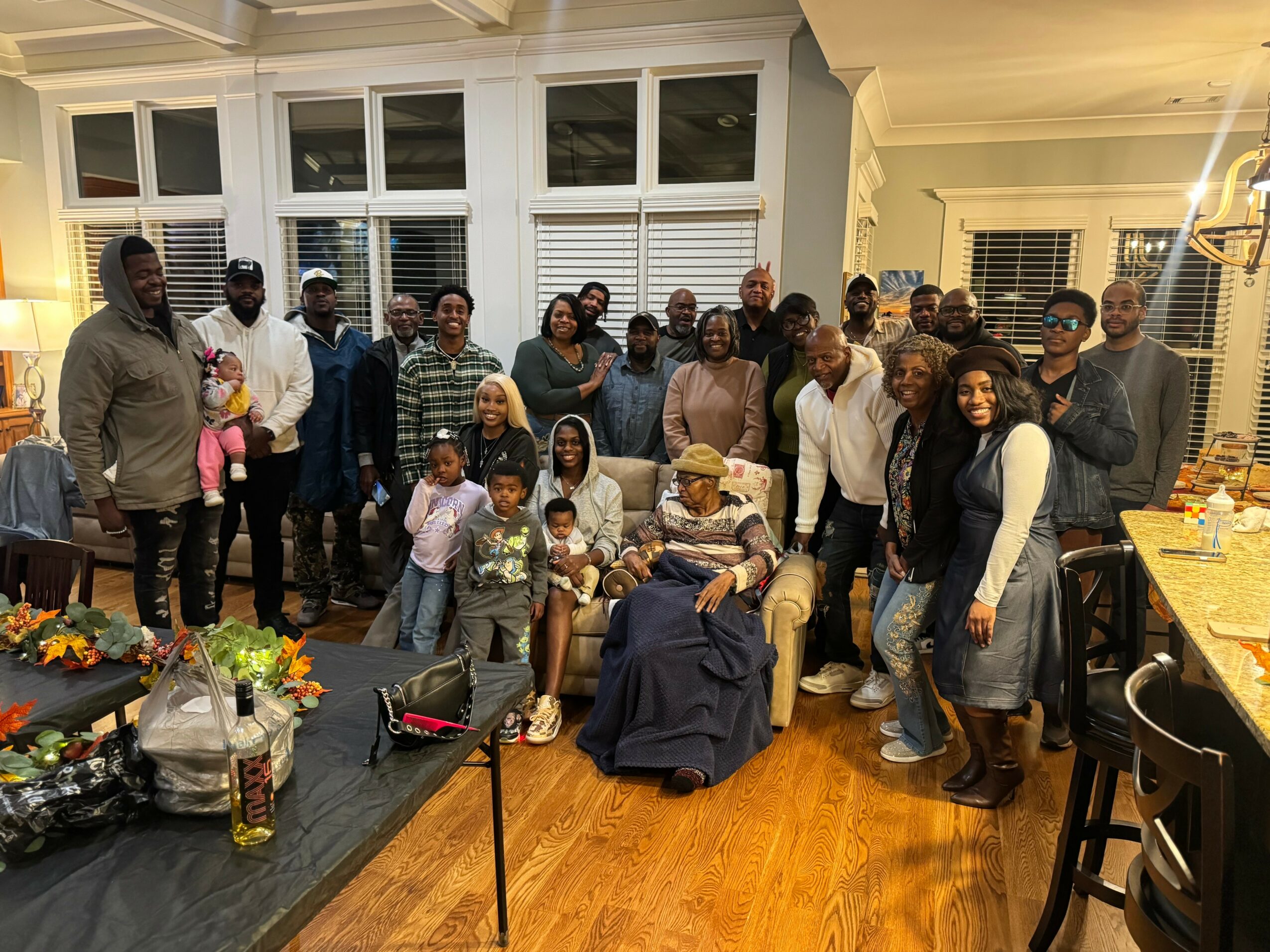 Group of people gathered in a living room, some sitting and some standing, with a woman in a wheelchair in the center.