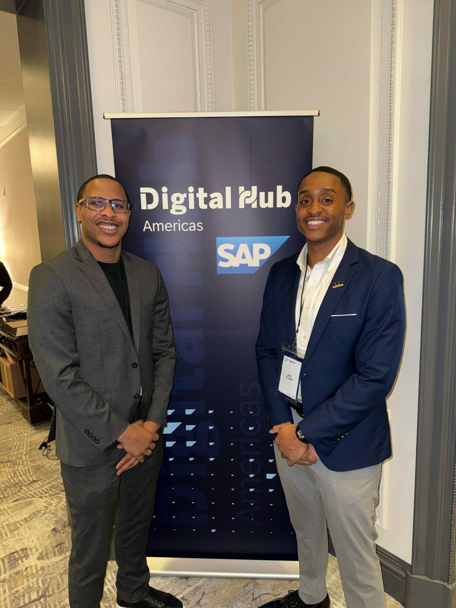 Two men in business attire standing in front of a SAP banner at a professional event.