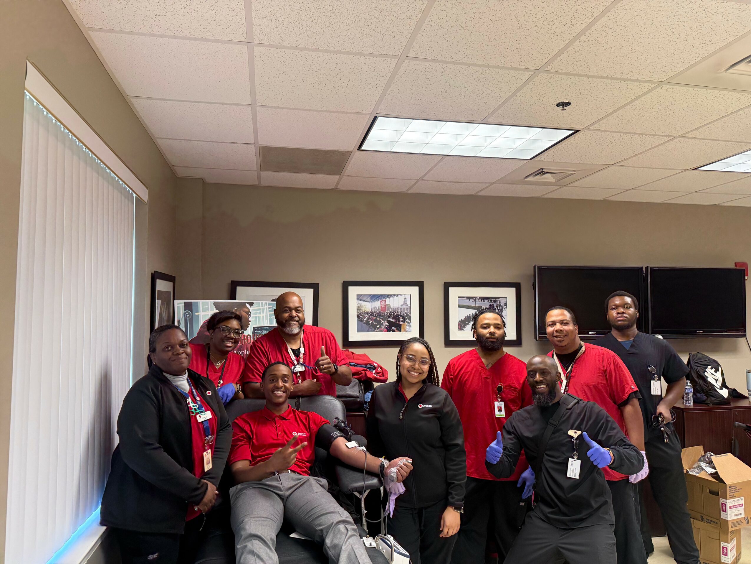 Group of eight people standing in an office room, some wearing red shirts, with framed pictures on the wall behind them.