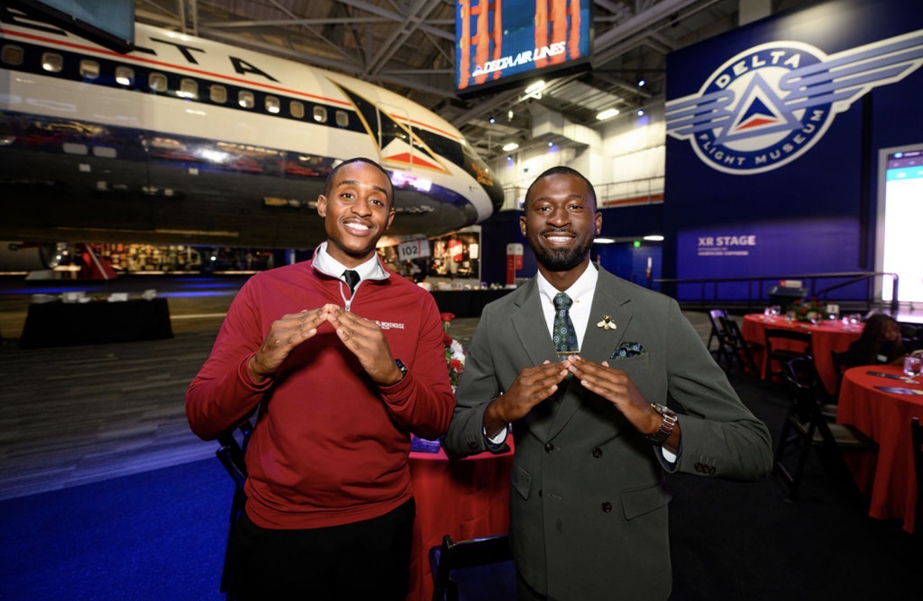 Two men in formal attire stand in front of a Delta airplane display at an indoor event, smiling and making hand gestures.