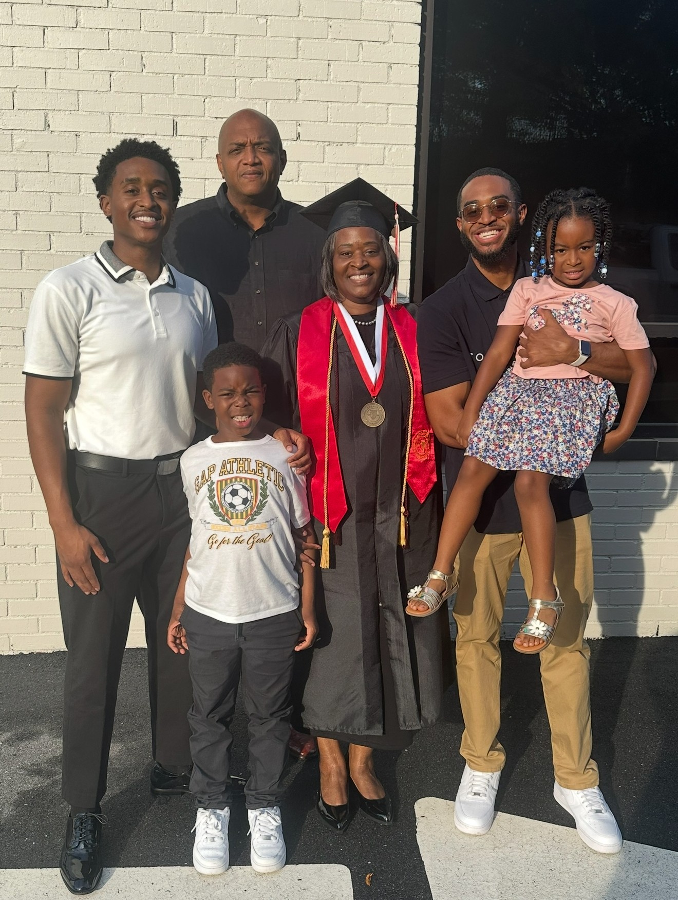 Group of six people, including a graduate in cap and gown with medals, standing outside near a brick wall and window.