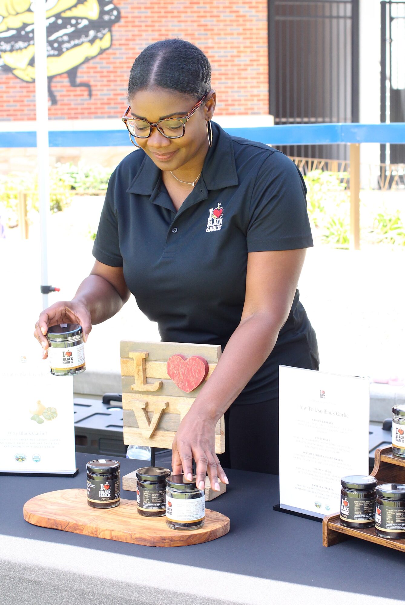 Woman with glasses and black hair in a ponytail, wearing a navy polo shirt, arranging jars on a table outdoors.