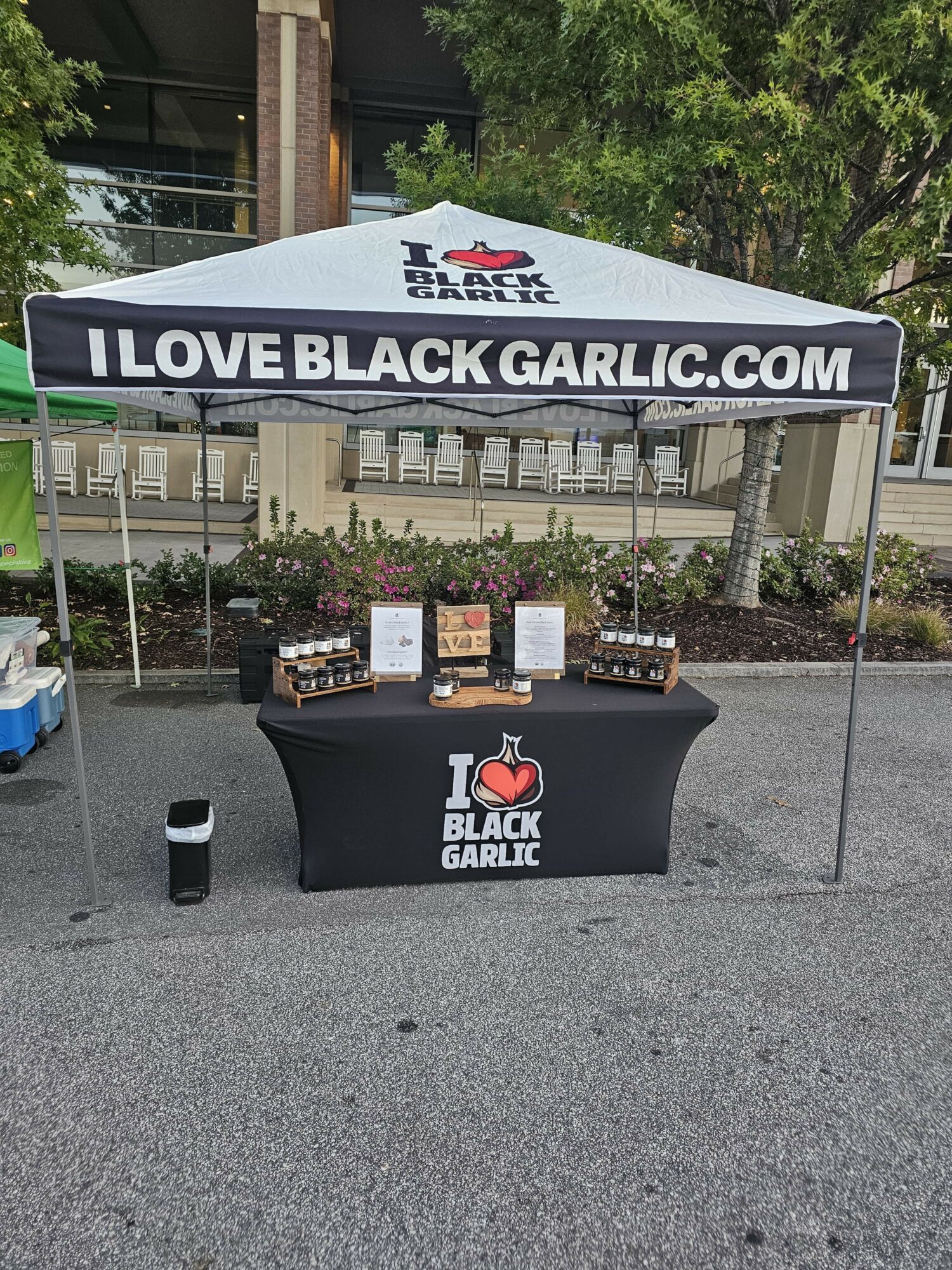 Outdoor booth with a black table and a canopy displaying 'I LOVE BLACK GARLIC' and website, set on a street with trees and buildings.