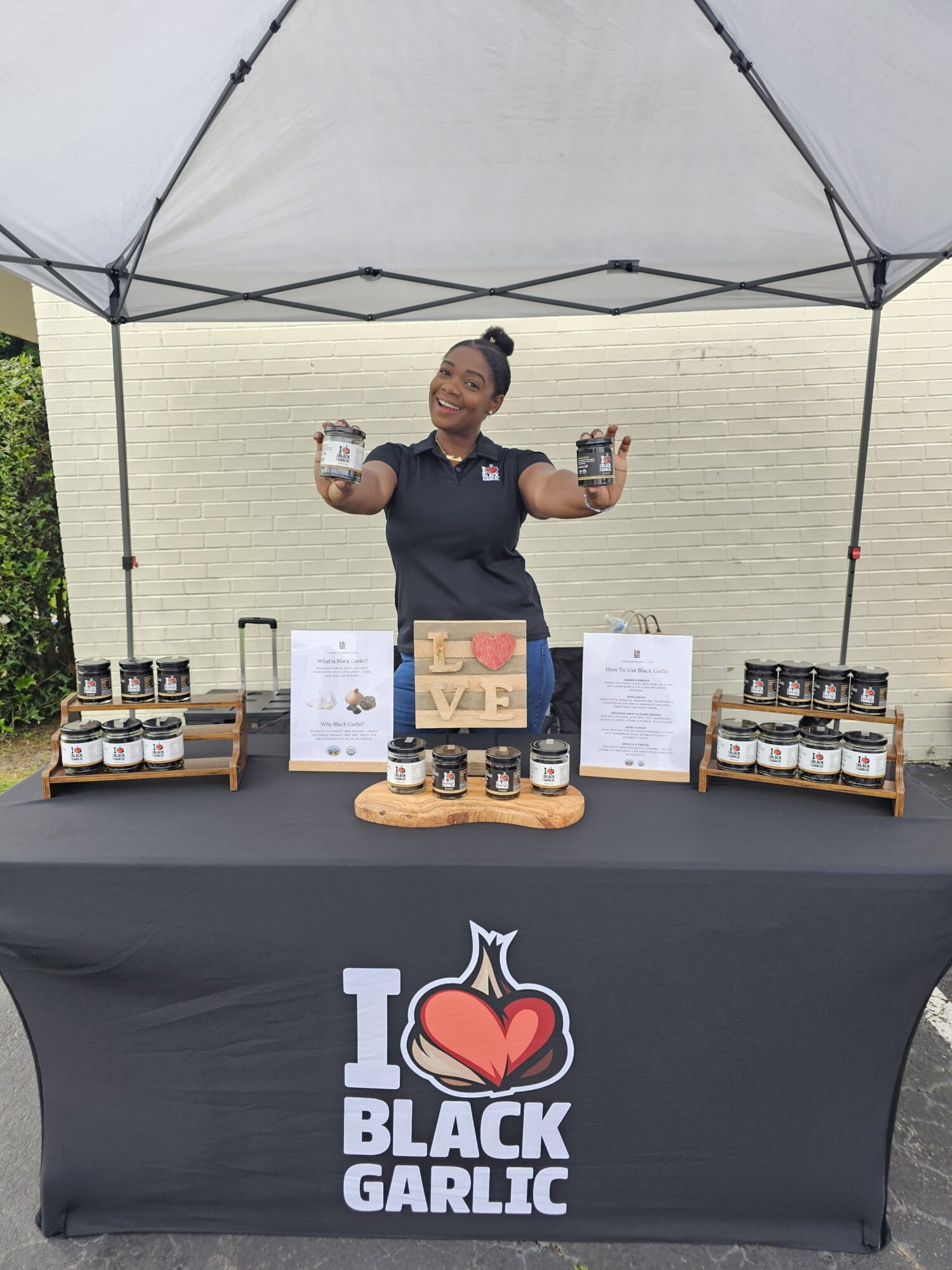 Person standing behind a table with jars, under a canopy, smiling, with a sign and display items, outdoors.
