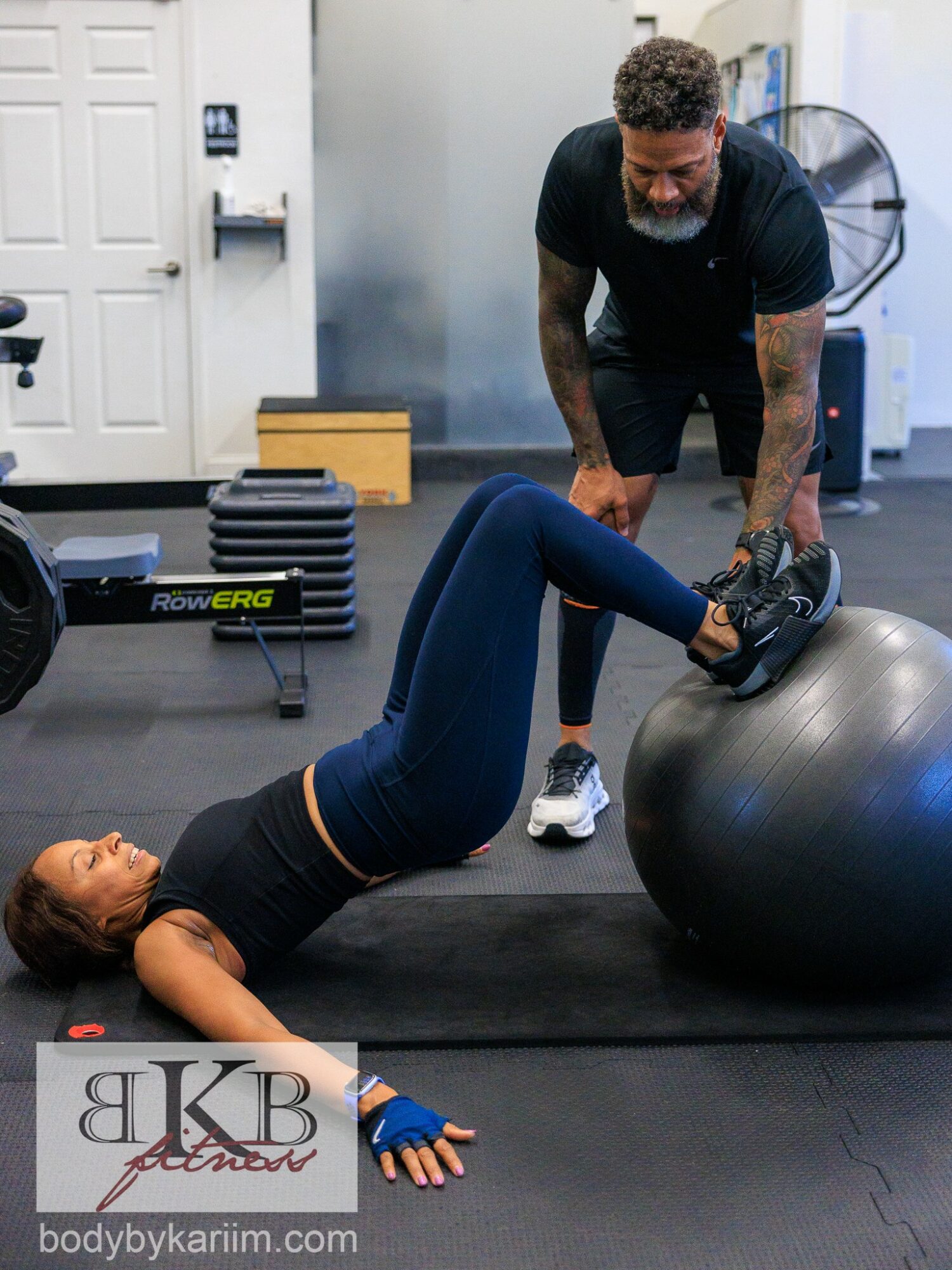 Woman lying on a mat lifting a weight with her legs, trainer assisting in gym setting.
