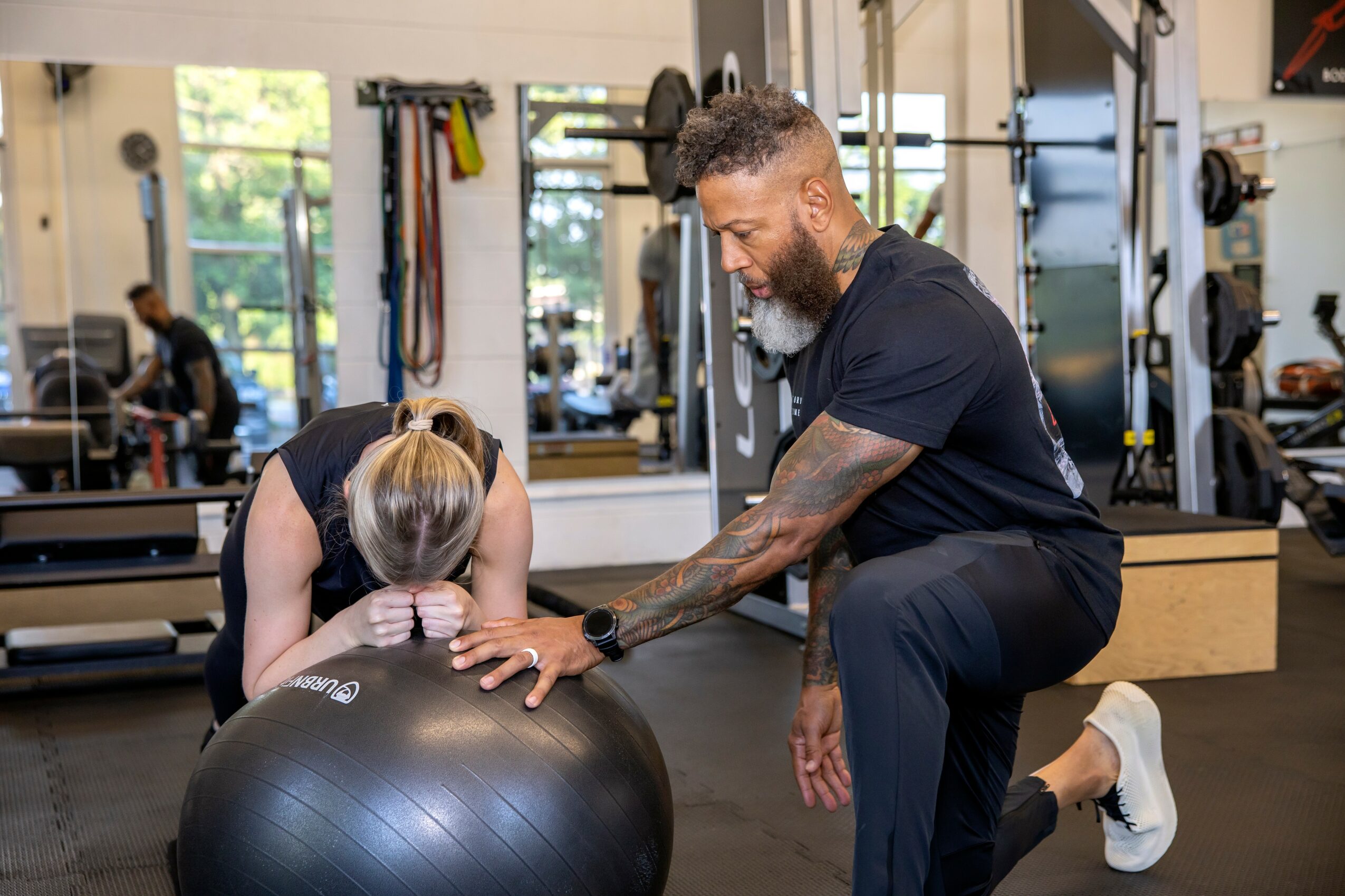 A woman and a trainer are in a gym, with the woman leaning on a large exercise ball, and the trainer assisting her.