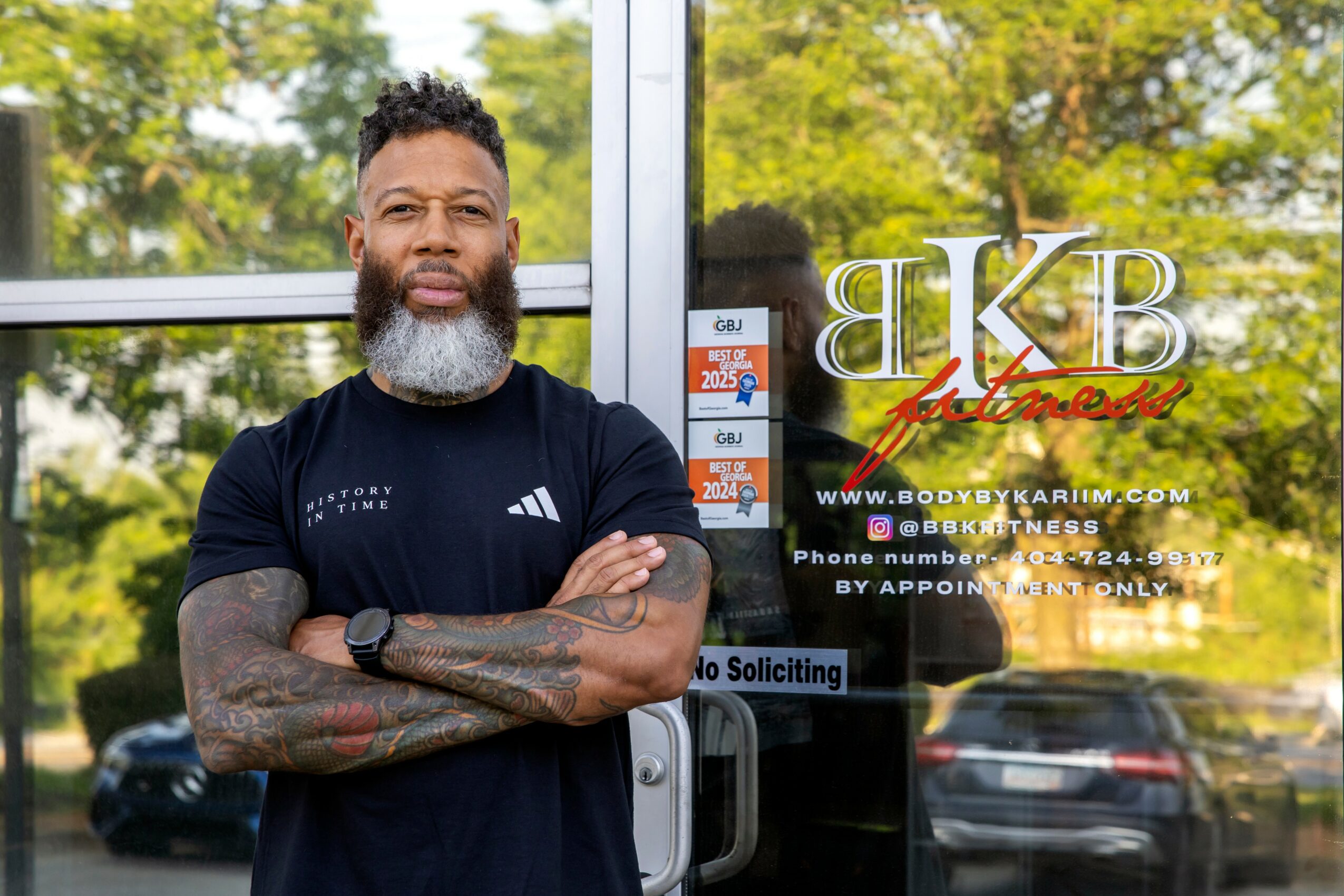 Bearded man with tattoos and athletic clothing stands outside in front of glass door with text and website, crossing arms.