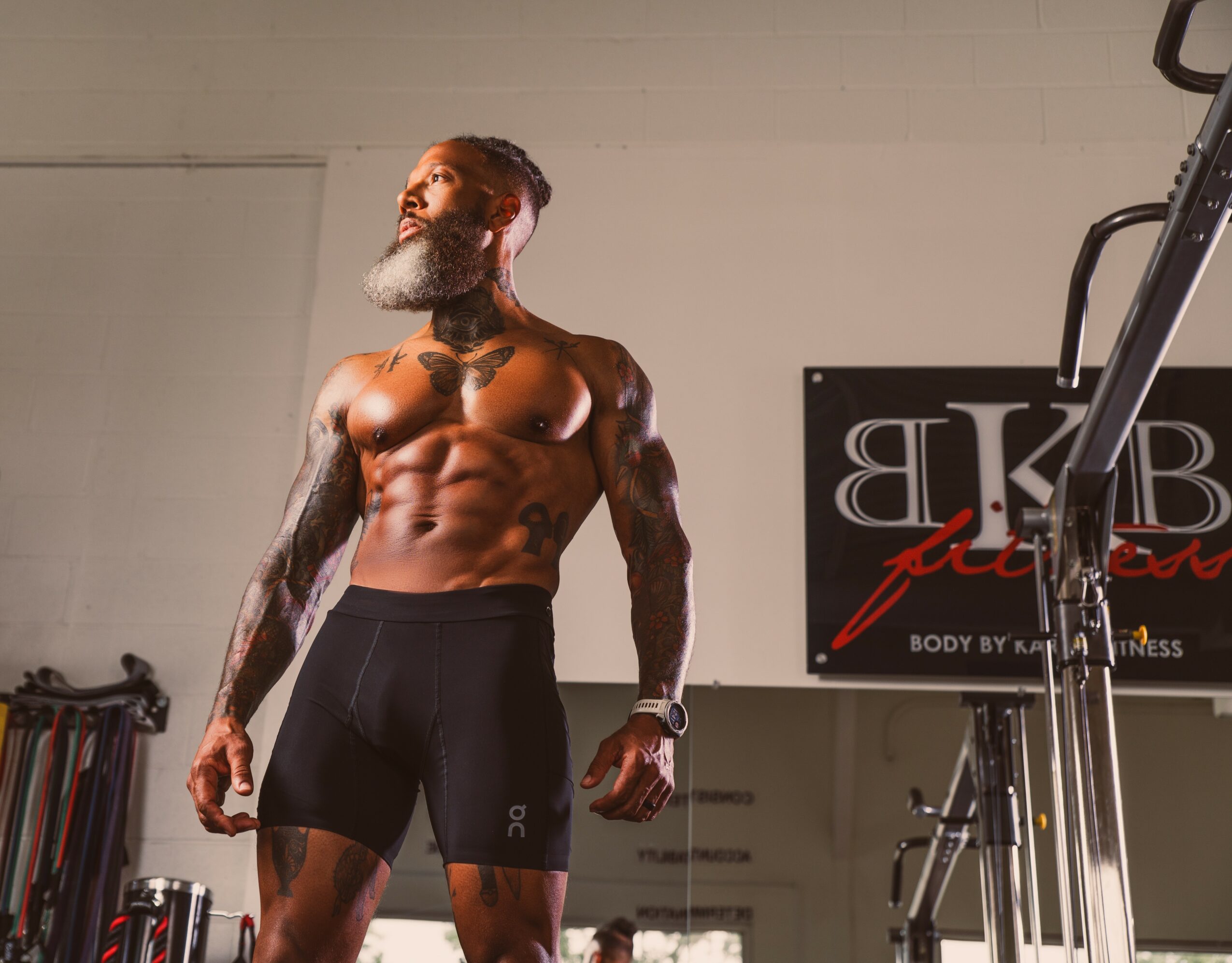 Muscular man with a beard and tattoos standing in a gym, looking upward, wearing black shorts, with gym equipment in background.